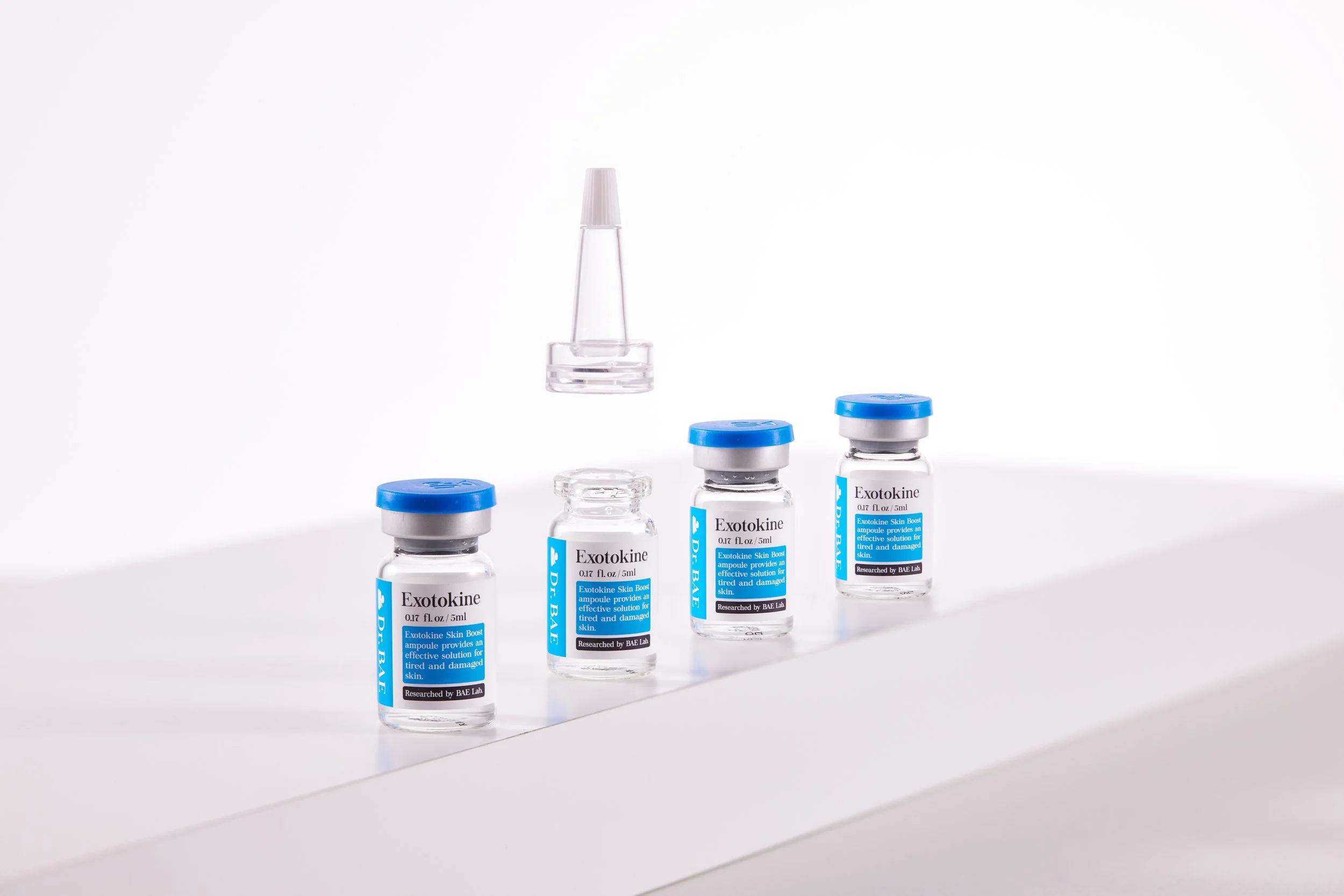 Four small glass vials labeled 'Exotokine' with blue and white labels, with two additional vials behind them, and a clear plastic applicator or dropper bottle floating above the vials against a plain white background.