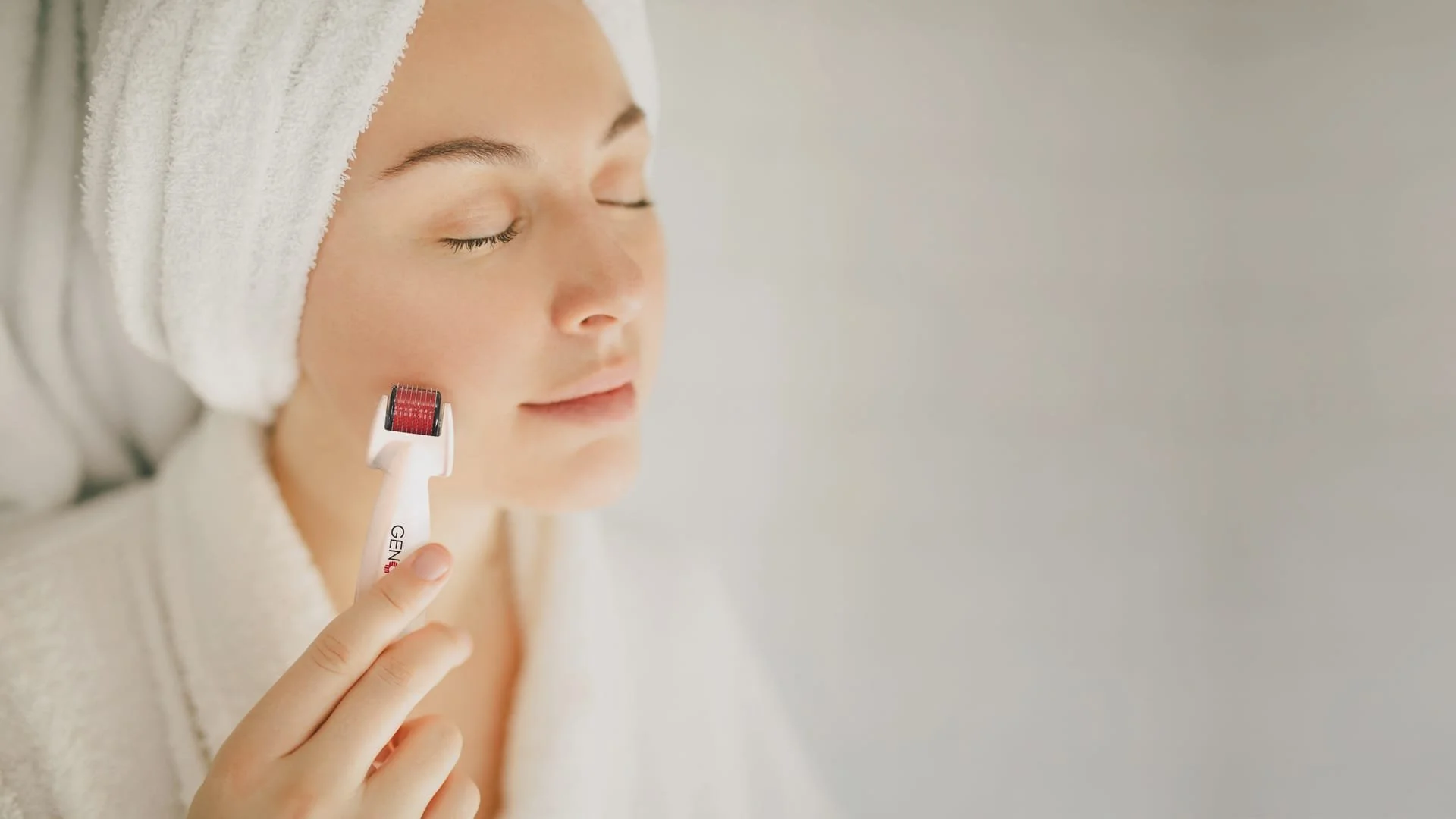 Woman in a towel with eyes closed using a facial roller on her cheek in a relaxing skincare routine.