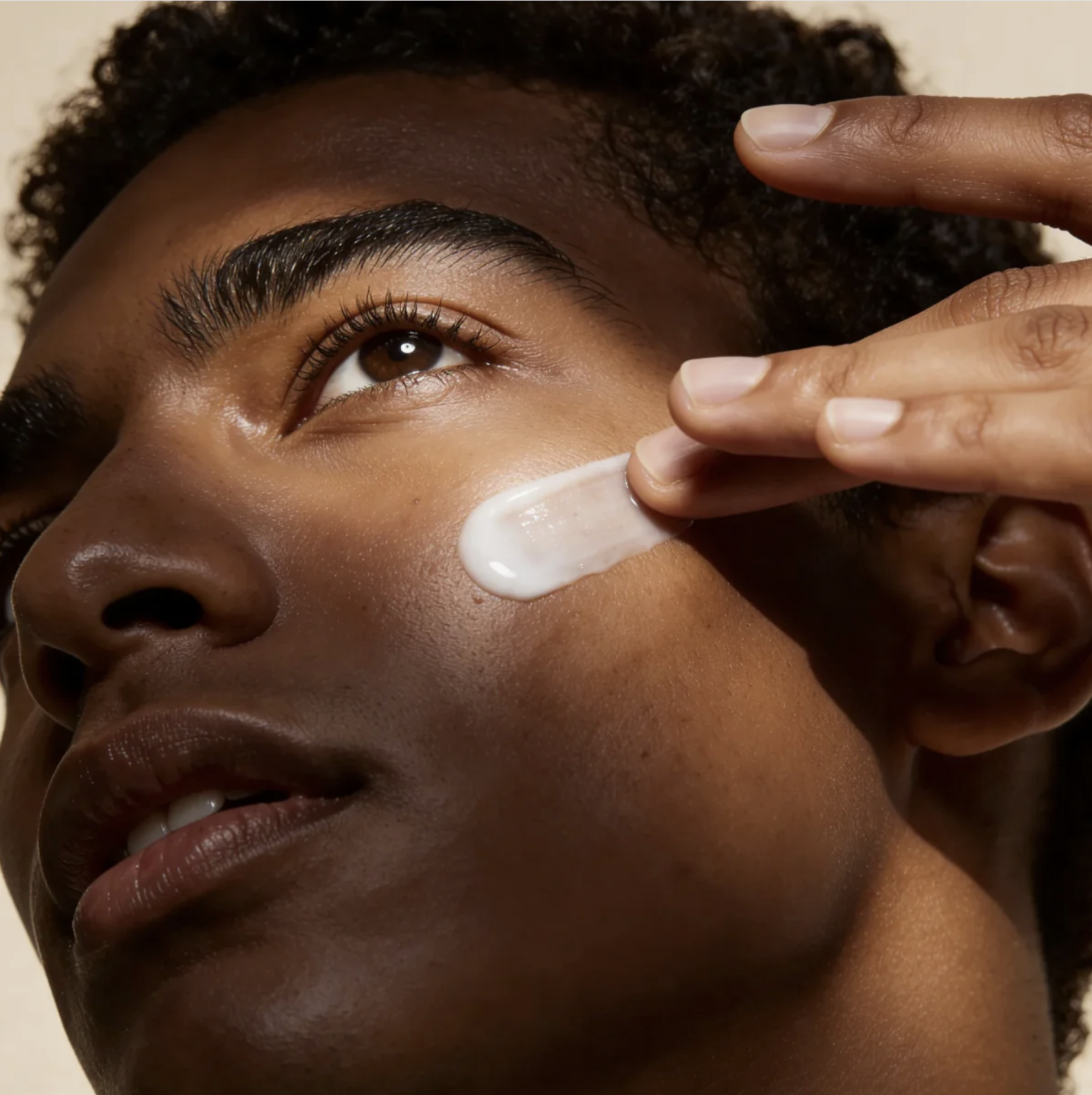 Close-up of a person with a smooth, dark complexion applying a white cream under their eye using a finger with neatly manicured nails.