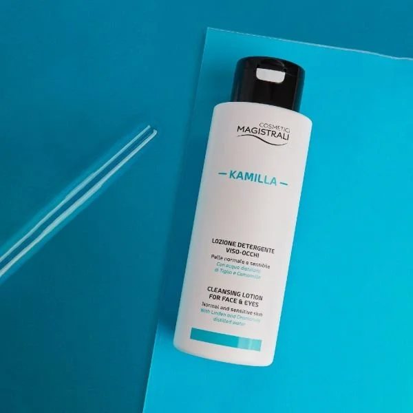 A bottle of Kamilla cleansing lotion for face and eyes on a blue background with a clear glass straw nearby.
