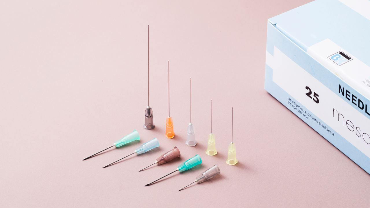 Assorted needles with colorful plastic hubs and a box of 25 sterile needles on a pink surface.