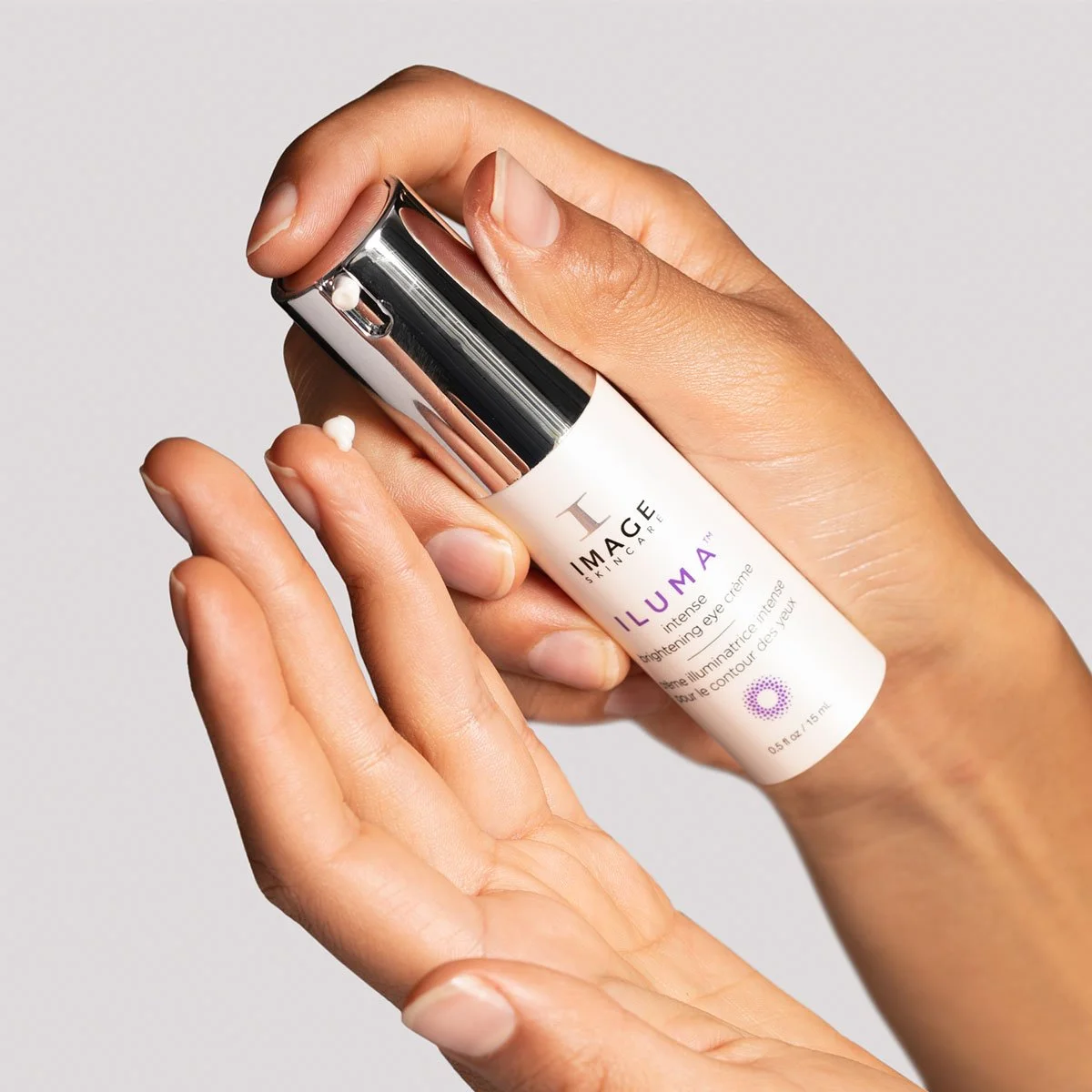 A person holding a white bottle of skincare product labeled 'Illuma' with a silver cap, and a few drops of cream on fingers.