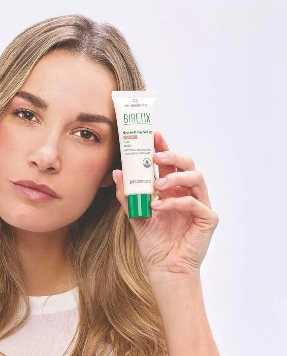 A woman with long, blonde hair holding a tube of Biretix Hydramat Day SPF30 moisturizer close to her face, against a plain white background.