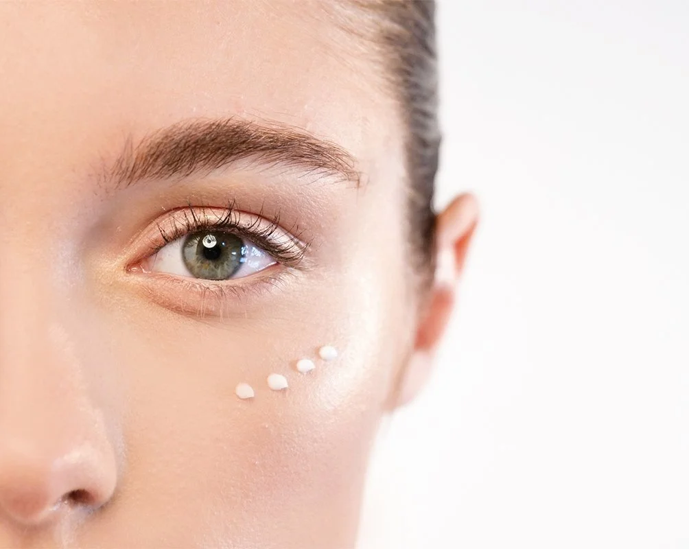 Close-up of a woman’s face showing her eye and cheek, with small dots of white cream on her cheek.