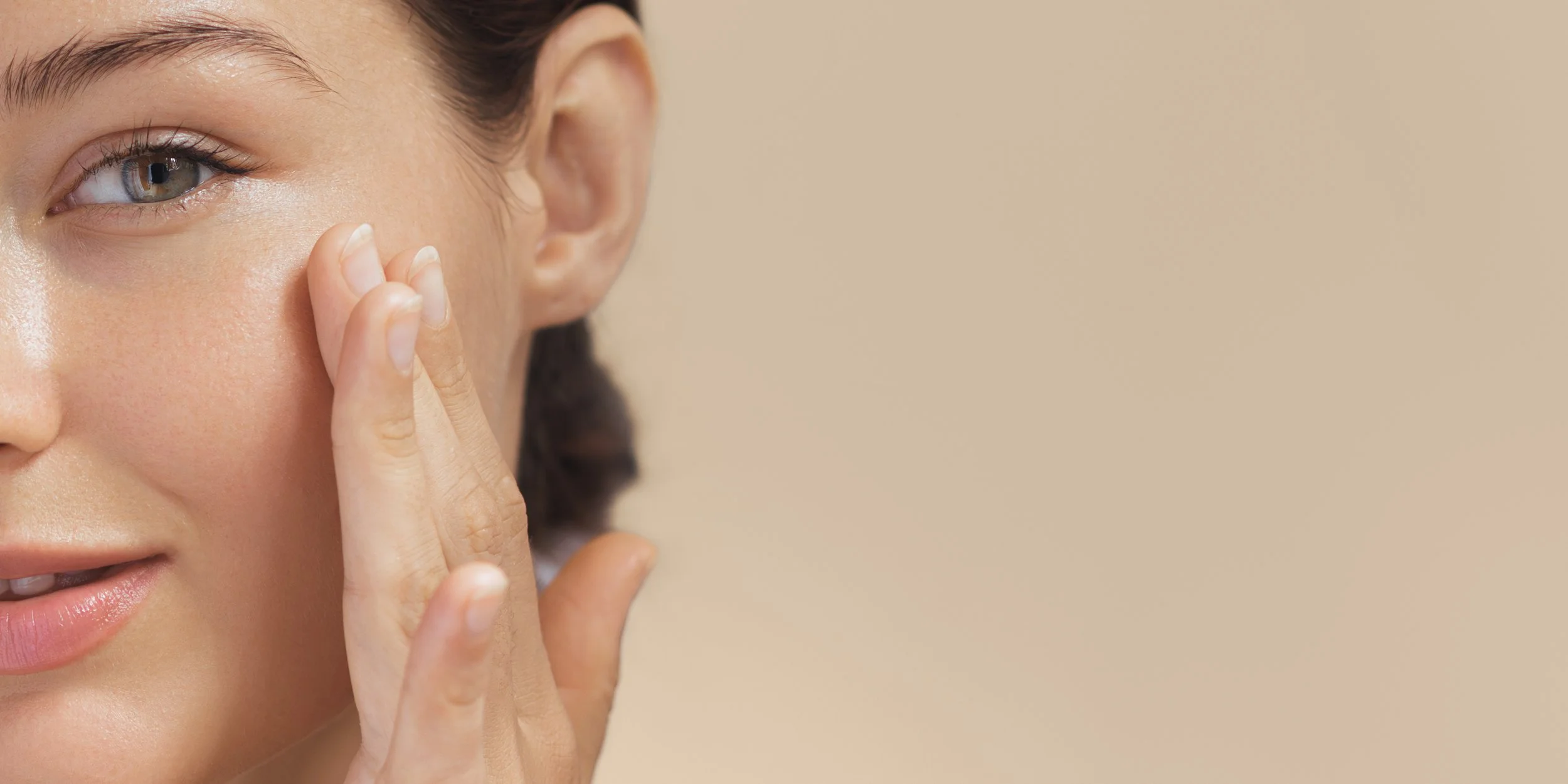Close-up of a woman with clear skin and blue eyes, gently touching her cheek with her index finger.