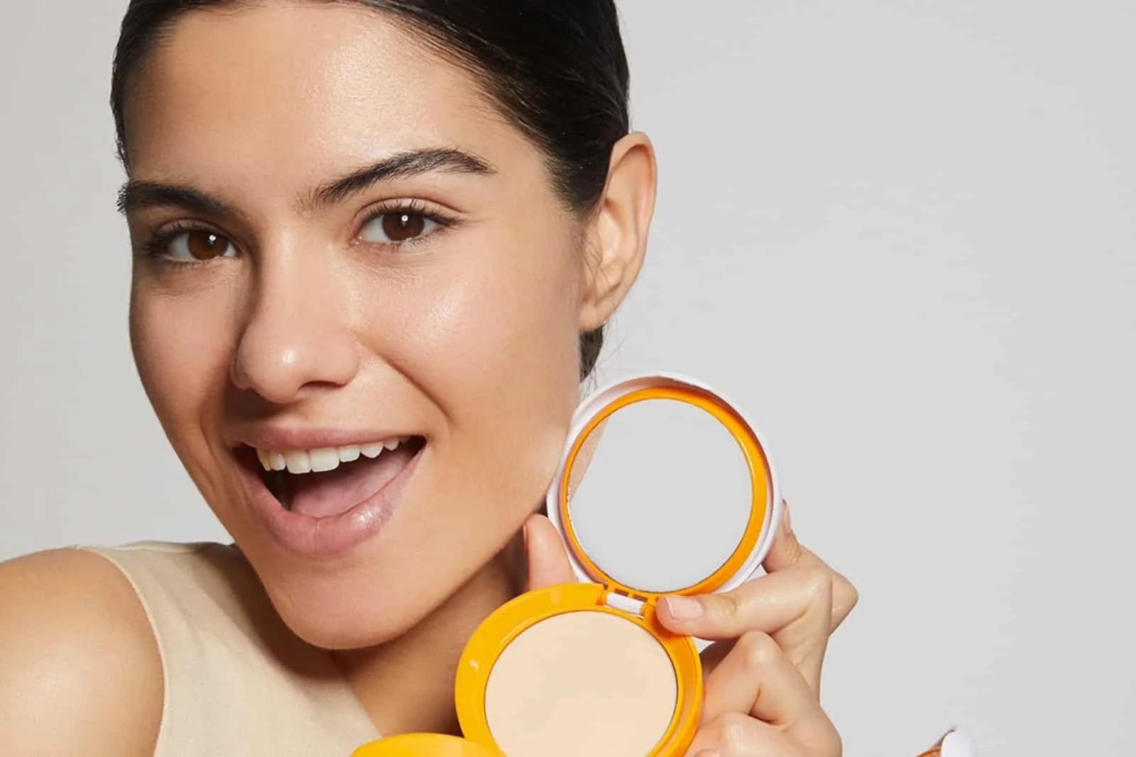 A woman with brown hair holds an orange compact mirror and lipstick, smiling with her mouth open against a plain light gray background.