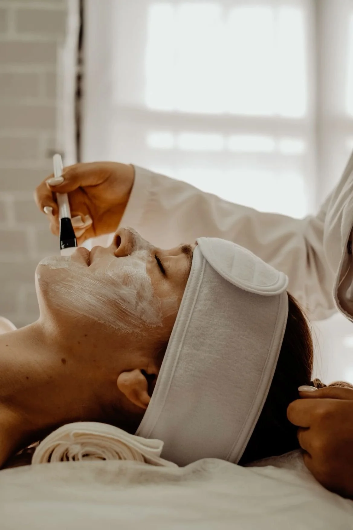 A woman lying down with a facial mask applied, receiving a facial treatment from a professional in a spa or skincare clinic.
