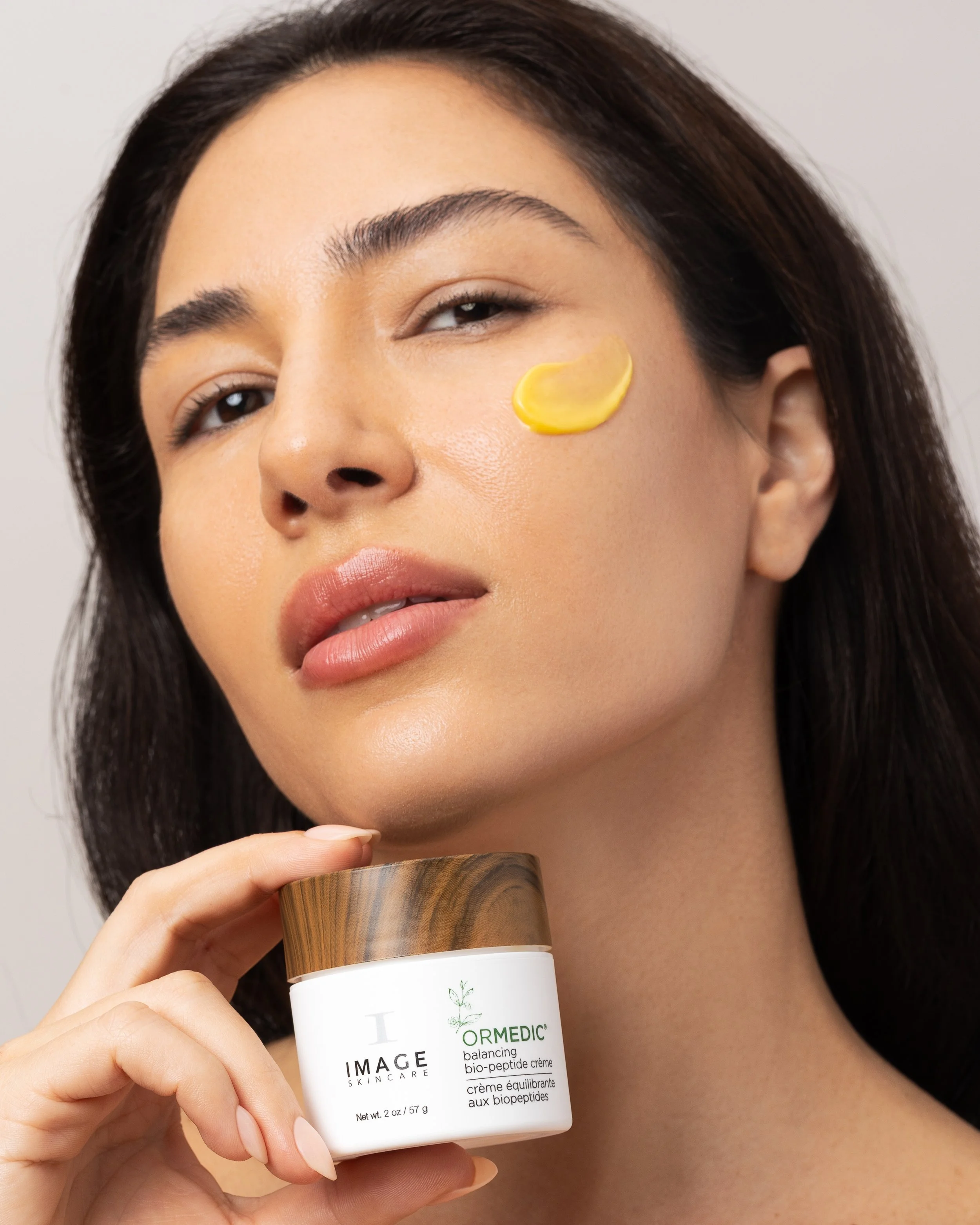 A woman with dark hair applying a skincare cream in a jar labeled 'ORMEDIC' to her face, holding the jar in her hand.