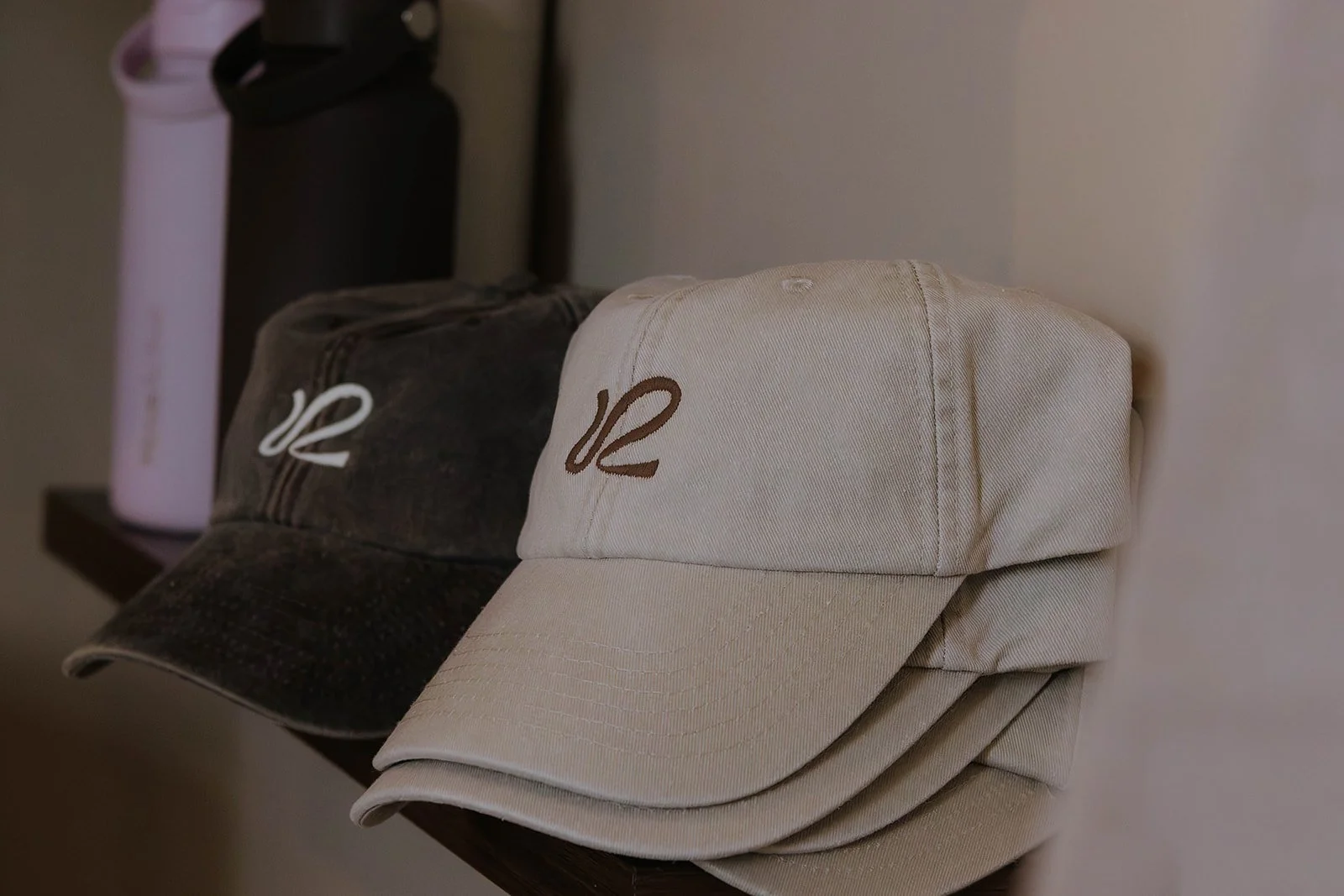 Three baseball caps are stacked on a wall shelf, with a white cap at the front, a black cap behind it, and a pink water bottle in the background.