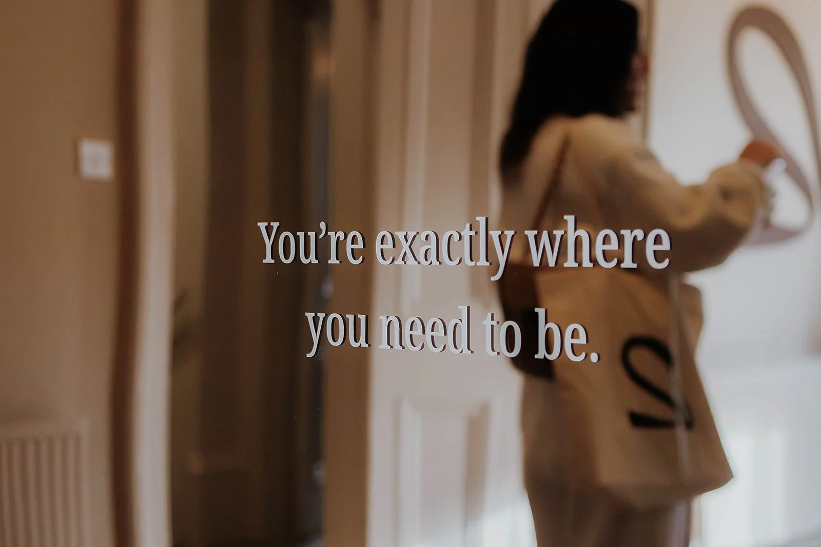 A woman in a beige trench coat with a bag over her shoulder looking at a mirror with a message that says, "You're exactly where you need to be."