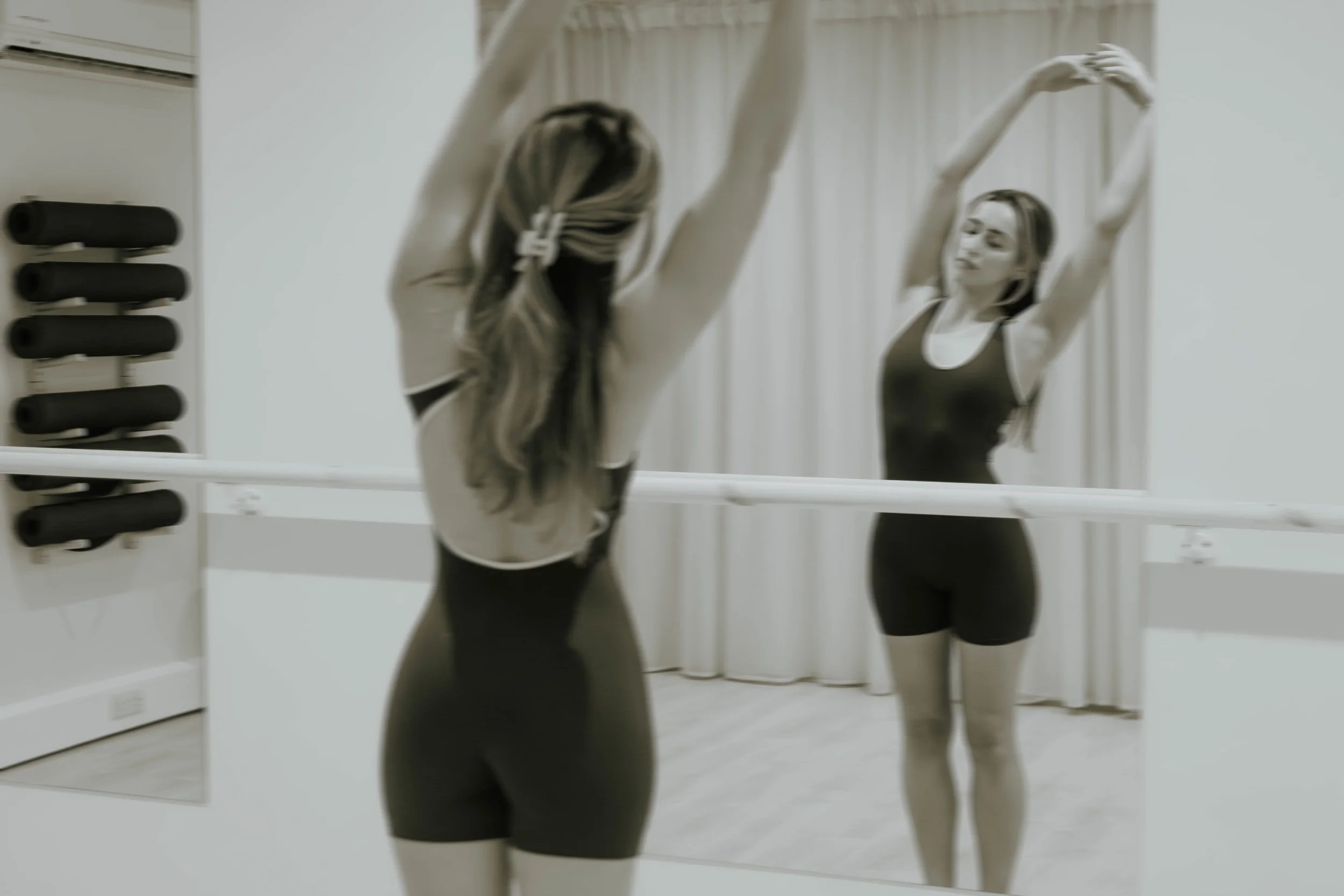 A woman in black workout clothes standing in front of a mirror with her arms raised, practicing a stretching or yoga pose in a fitness room.