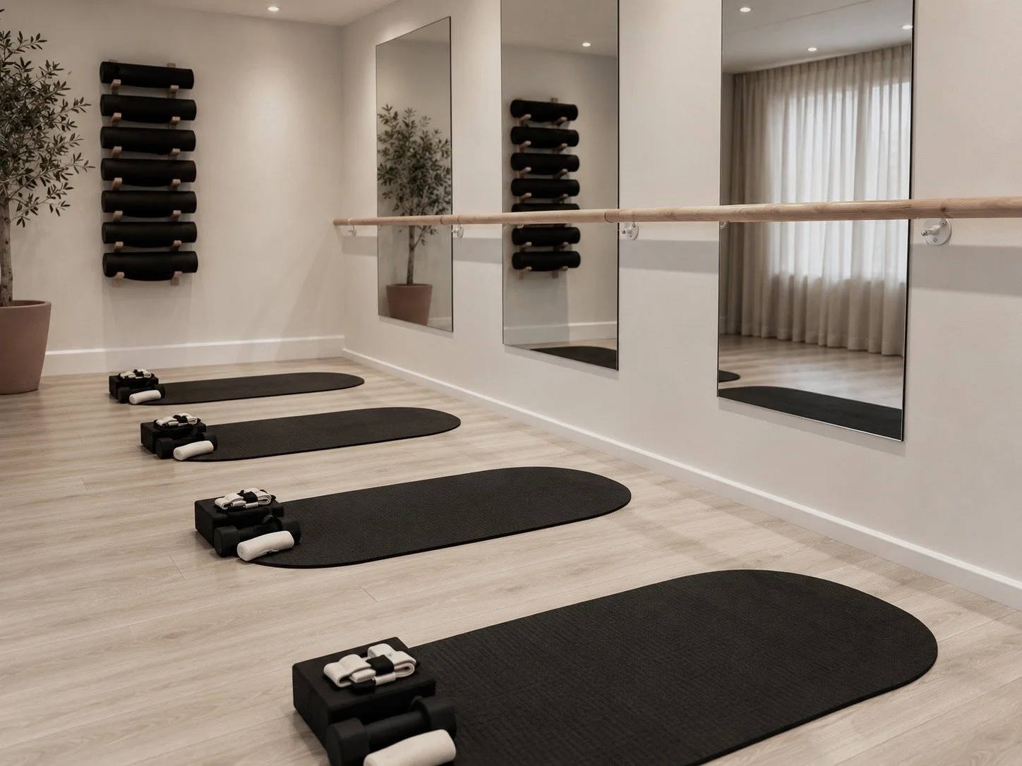 Indoor fitness studio with four black yoga mats and pilates reformer machines, large mirror, wall-mounted black foam rollers, potted plant, and wood flooring.