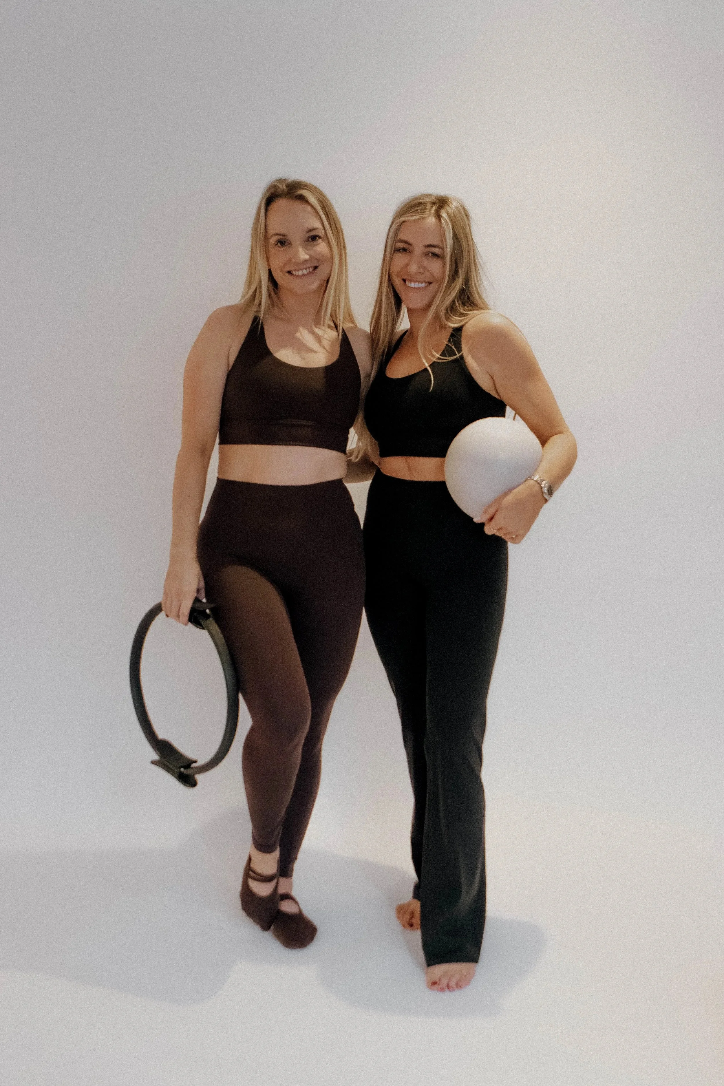 Two women in black athletic wear standing against a plain white background, smiling at the camera, with one holding a yoga block and the other holding a white ball.