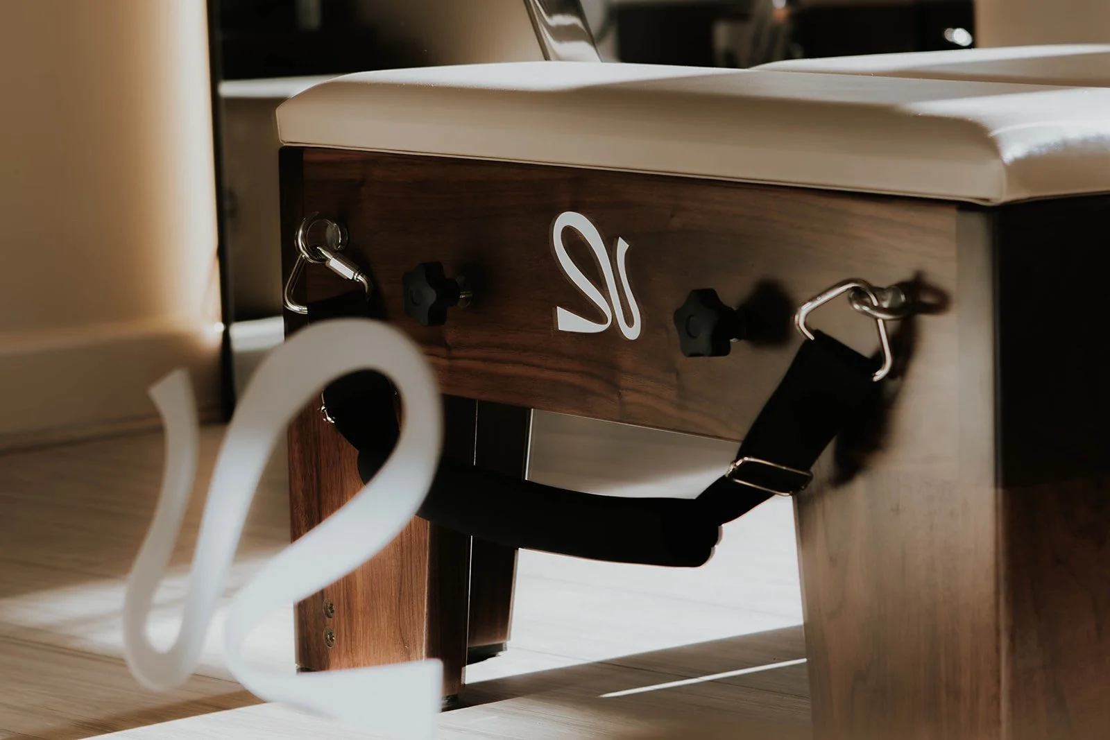 Close-up of a paddleboard with a wooden finish, white logo, and attached black padded foot straps, resting indoors.