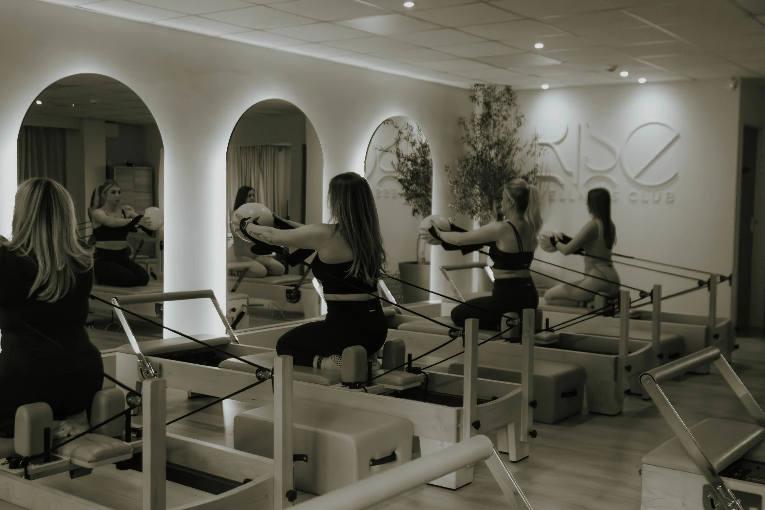 A fitness class in a gym, with women using exercise machines, with illuminated arches and a wall with a logo in the background.