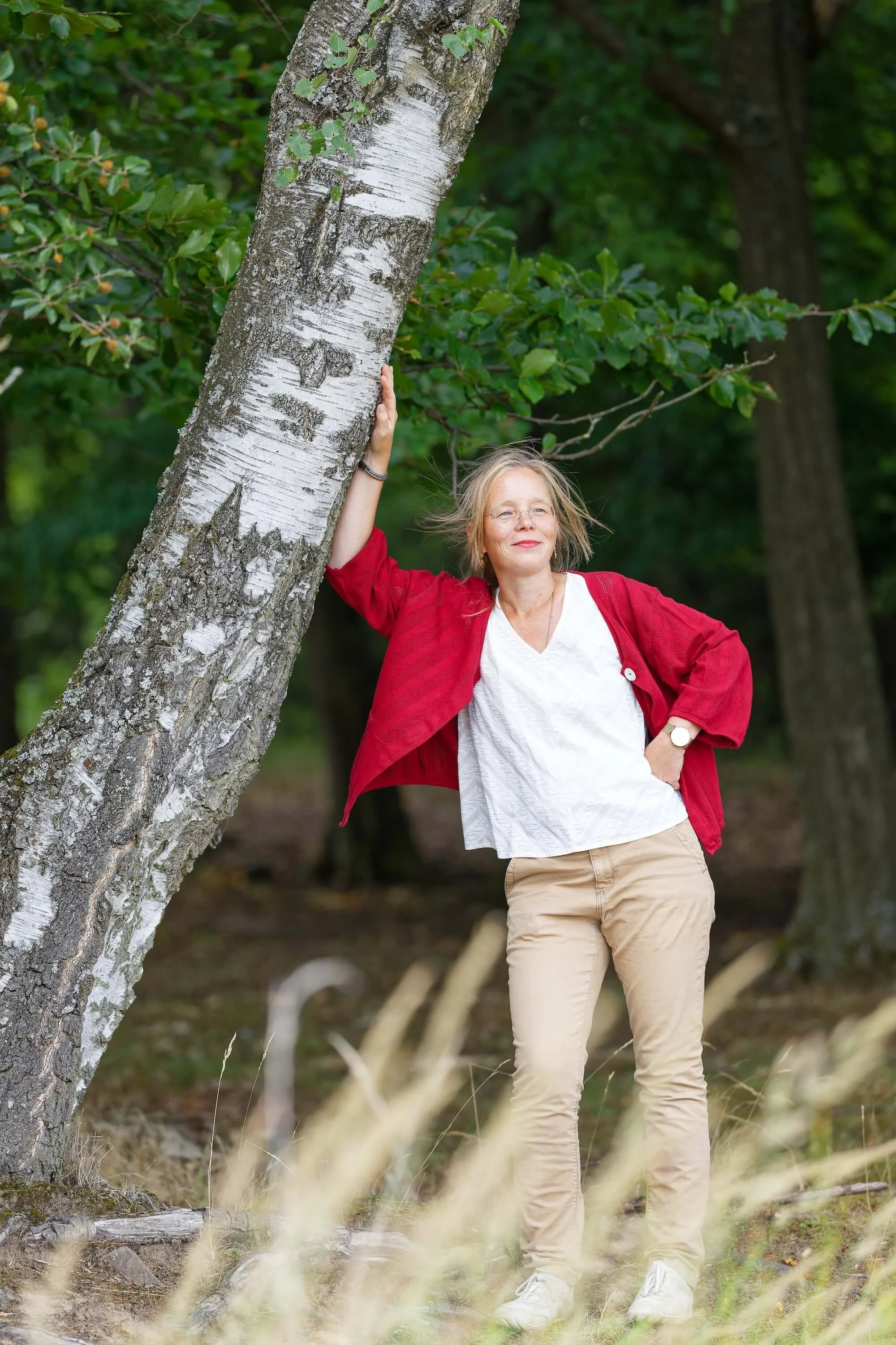 Eine Frau mit Brille, rotem Cardigan, weißem T-Shirt und beiger Hose, steht im Wald neben einem Baum, lächelt und berührt den Baum mit der Hand.