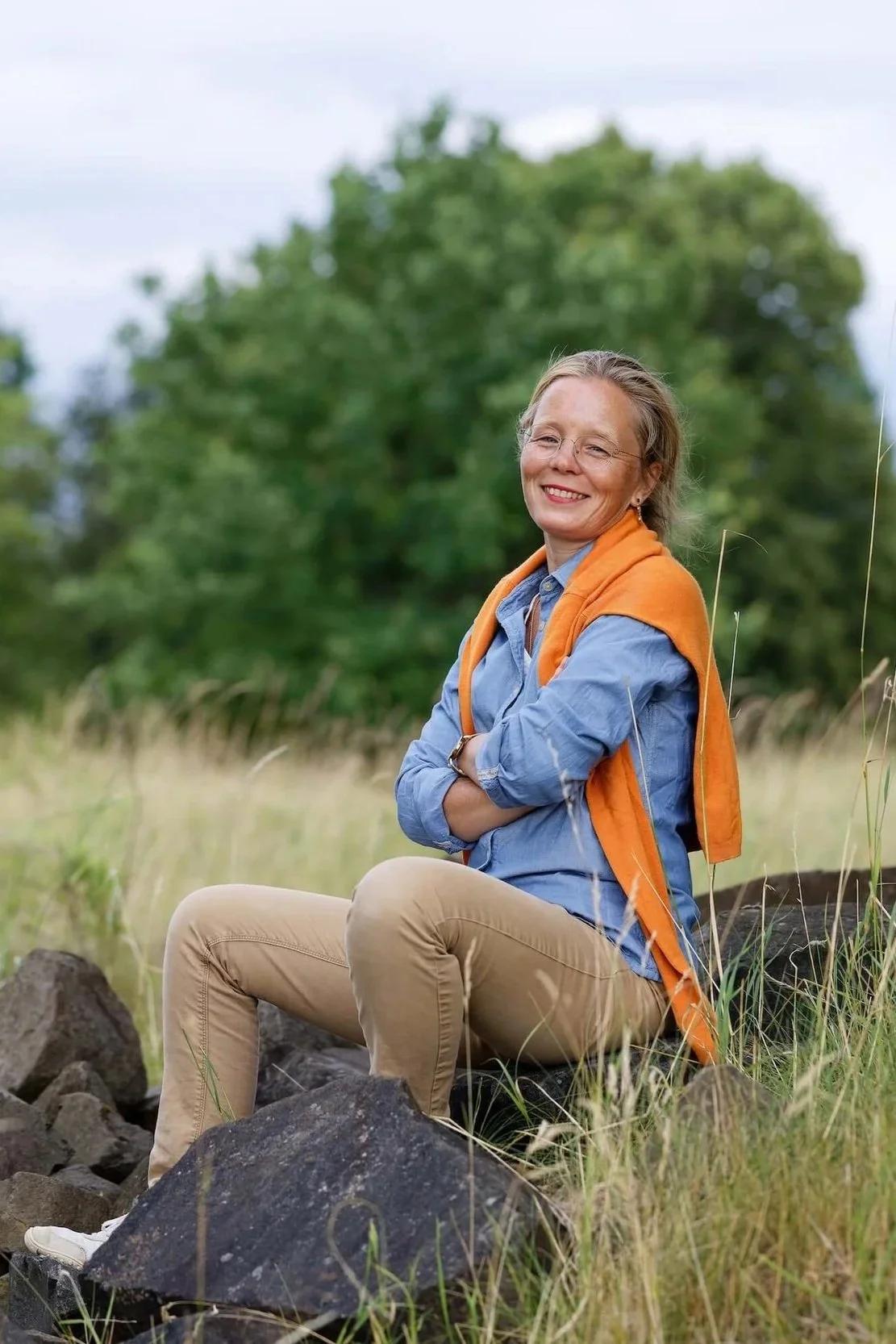 Eine lachende Frau mit Brille sitzt auf Steinen in einem Feld, im Hintergrund Bäume, trägt eine blaue Bluse, einen orangen Pullover um die Schultern, beige Hosen und weiße Schuhe.