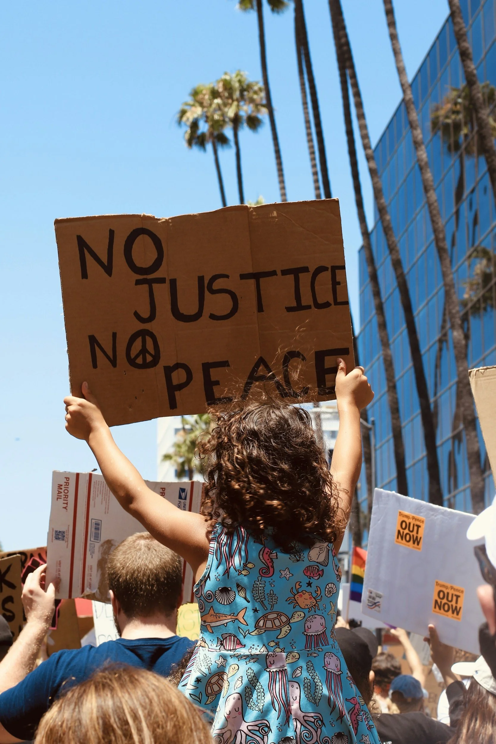 A child with curly hair holding a protest sign at a rally or protest in a city with palm trees and modern glass buildings. Civic engagement is crucial.