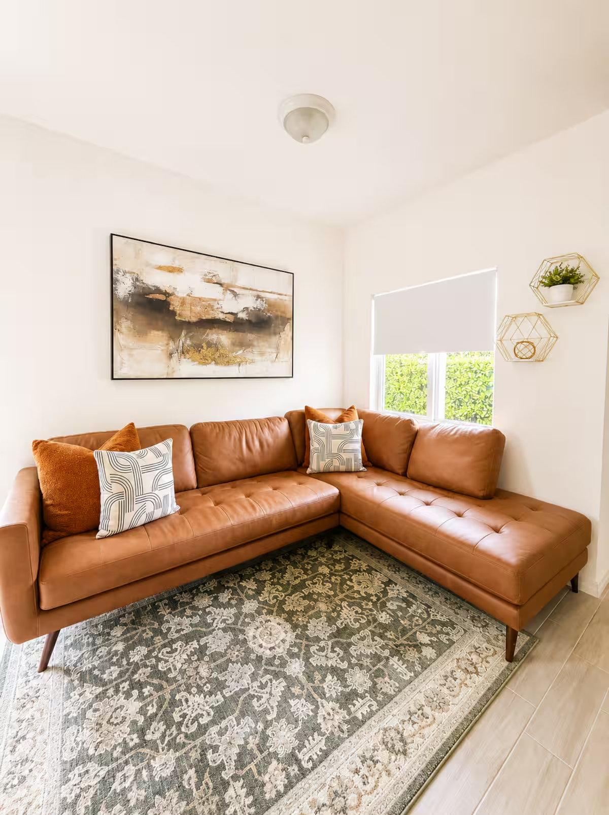 Living room with a brown leather sectional sofa, decorative pillows, a large abstract painting on the wall, and two gold hexagon-shaped wall shelves holding a small plant and a decorative object. A window with a white blind lets in natural light, and there is a patterned area rug on the floor.