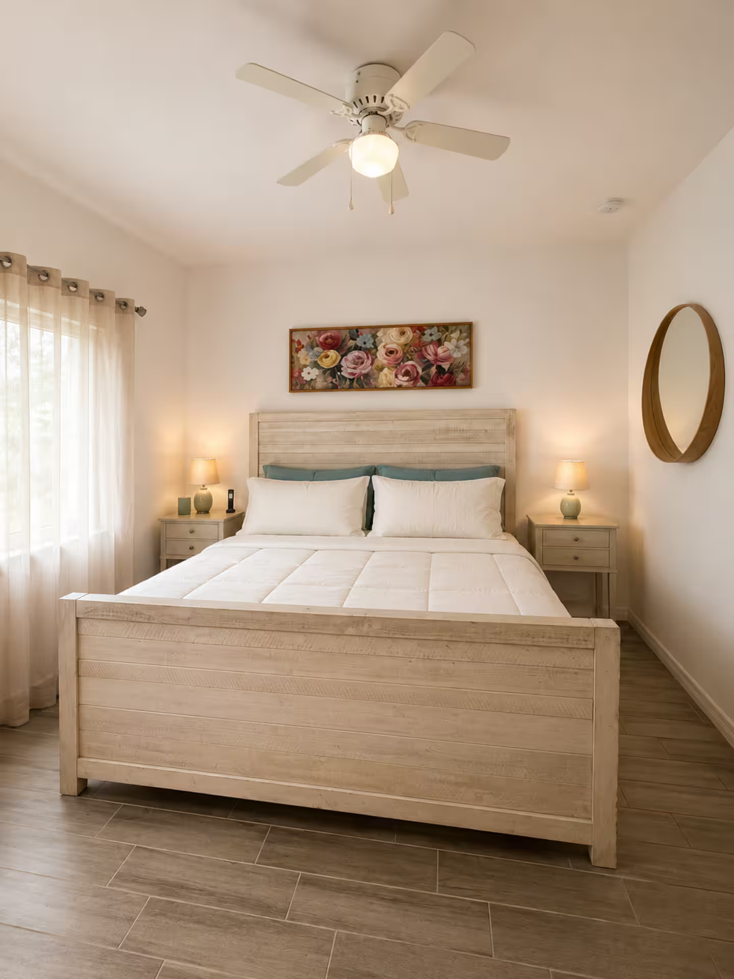 A bedroom with a white wooden bed frame, white bedding, and four pillows. There are two nightstands with lamps on each side of the bed, and a framed floral painting above the headboard. Light curtains cover a window on the left, and a round mirror hangs on the right wall. The ceiling has a white fan with a light.
