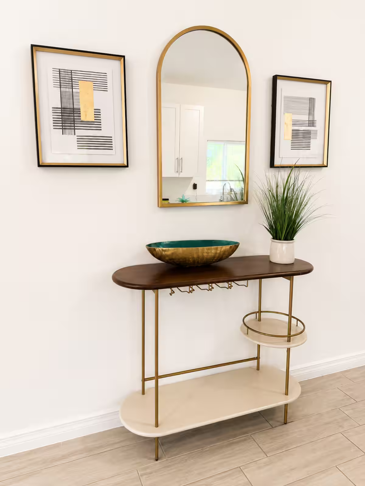 A beige and dark wood console table with gold accents, a green decorative bowl, and a potted plant with tall green grass in a white pot. Above the table is an arched mirror flanked by two abstract black and white framed art pieces on a white wall.