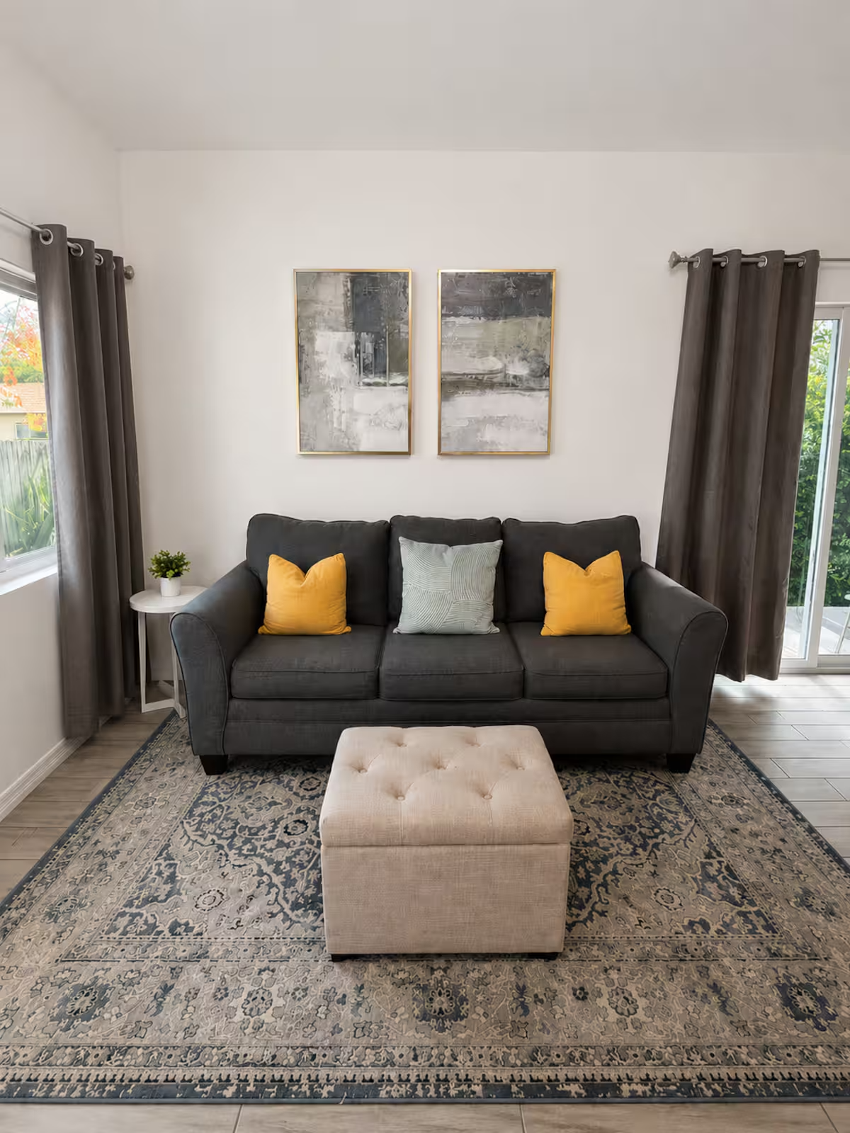 Living room with a dark gray sofa with three cushions, two yellow and one light gray, on a decorative area rug. There are two abstract art pieces hanging above the sofa. To the left, a side table holds a small potted plant. Dark gray curtains frame a window on the left, and a sliding glass door with similar curtains is on the right, letting in natural light.