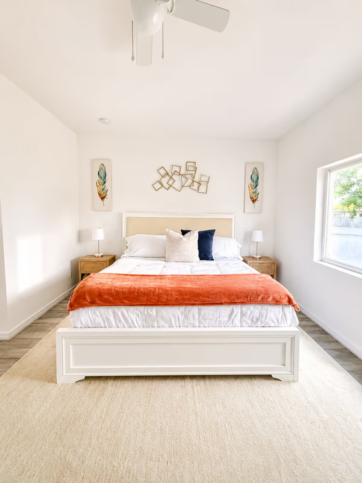 Bright bedroom with a white bed, orange throw blanket, two nightstands with lamps, and wall art featuring feathers.