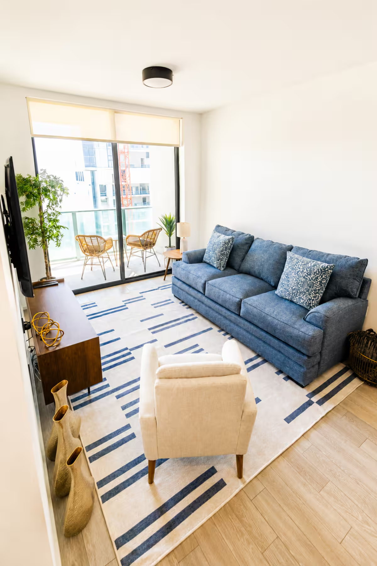 Living room with blue sofa and pillows, white armchair, wooden console table with decorative items, flat-screen TV, glass sliding door leading to balcony with two wicker chairs, potted plants, area rug with blue and white geometric pattern, wooden flooring, and ceiling light fixture.