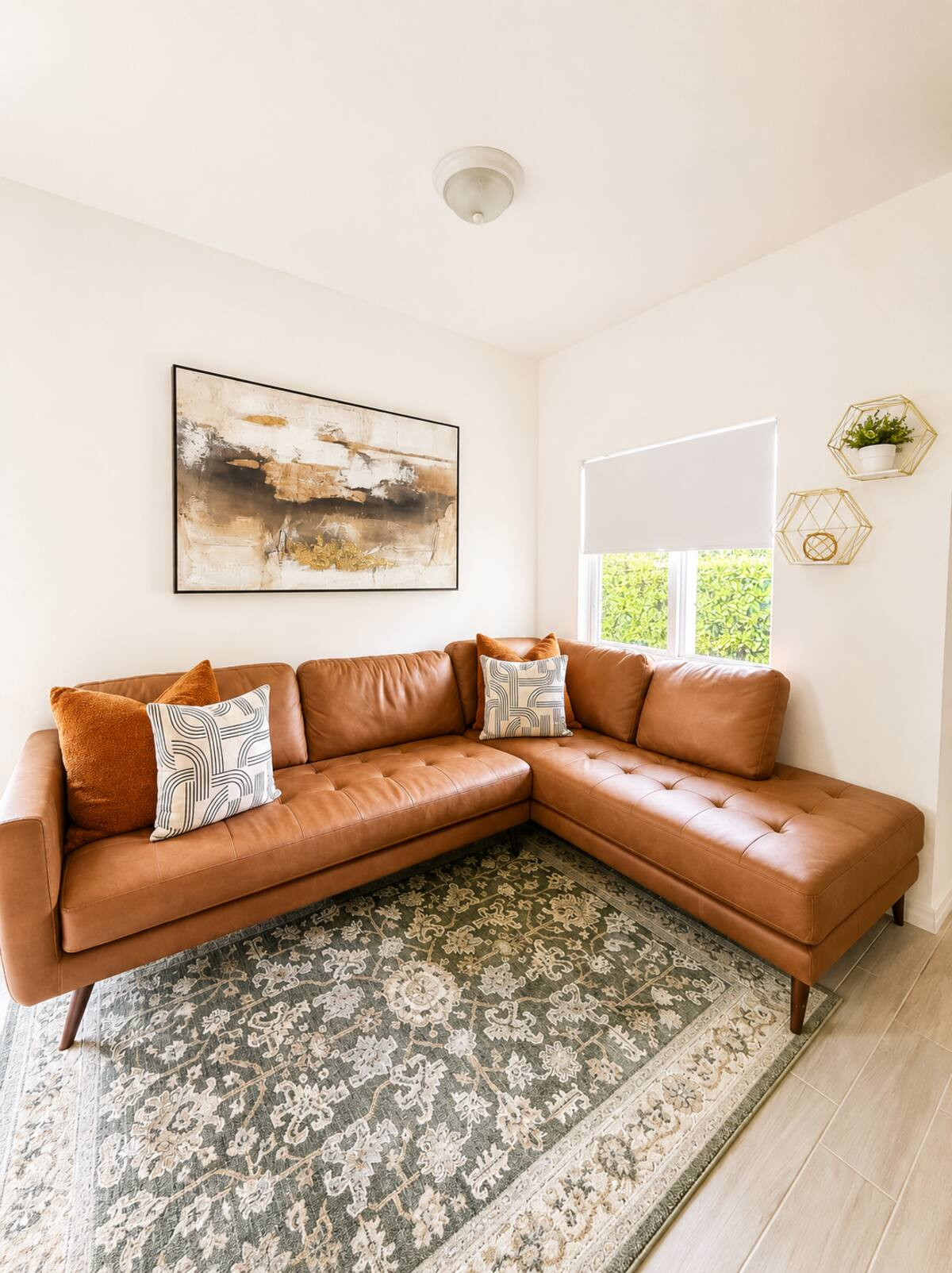 Living room with a tan leather sectional sofa, decorative pillows, an abstract wall art piece, a window with sheer valance, and geometric wall shelves with plants.