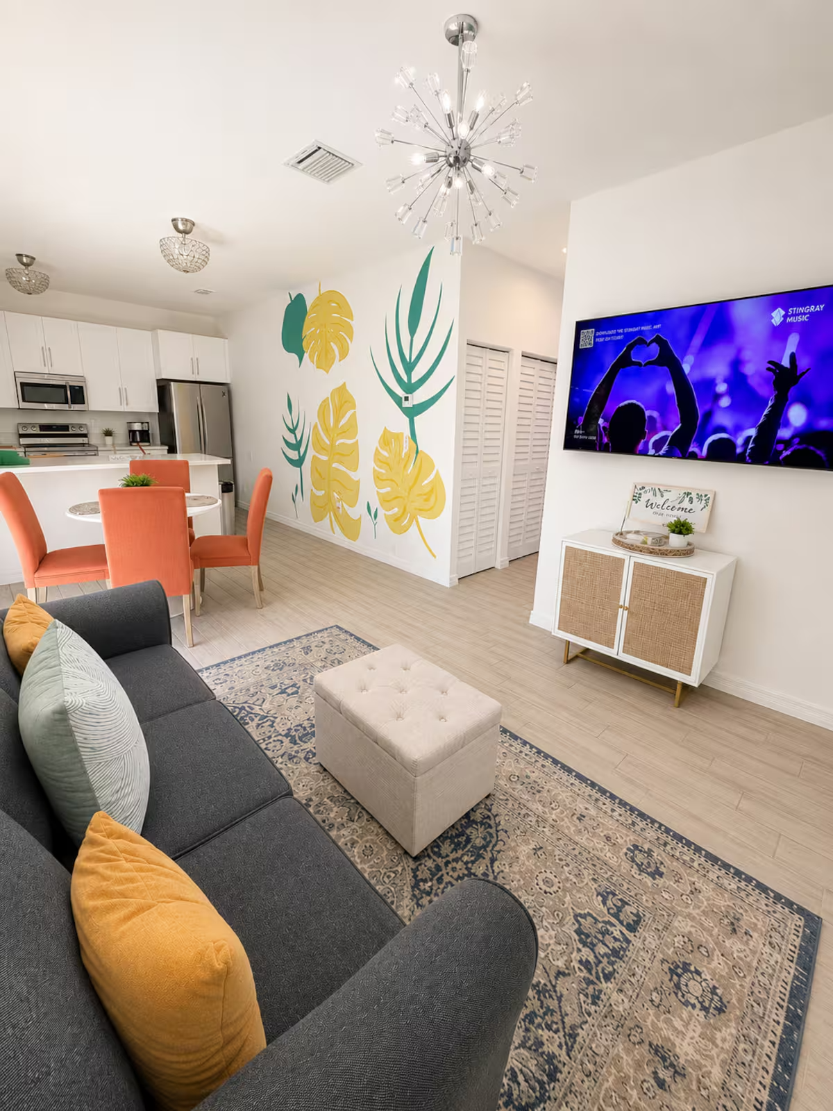 Modern living room with a black sofa decorated with yellow and light green pillows, a beige tufted ottoman, a patterned area rug, and a large TV mounted on the wall. The room features a white cabinet with a welcome sign, a colorful wall mural of tropical leaves, and a dining area with coral-colored chairs and a round table. The kitchen with white cabinets and stainless steel appliances is in the background. A contemporary chandelier hangs from the ceiling.