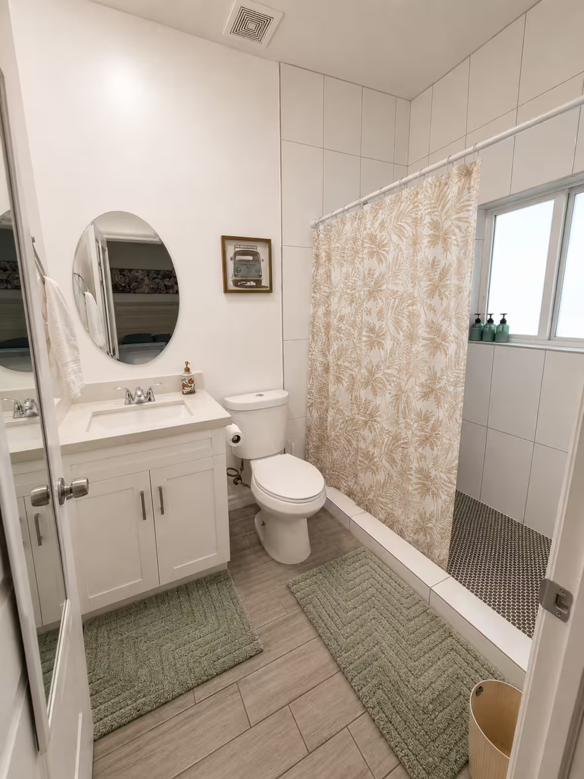 A small bathroom with white walls and a dark wood floor. It has a white vanity with a sink and an oval mirror. A toilet is next to the vanity, and a shower area with a beige leafy-patterned curtain is to the right. On the windowsill, there are three green soap dispensers. Two gray rugs are on the floor, and there's a framed picture of a van on the wall.