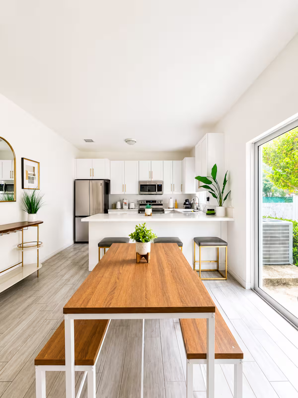 Bright, modern kitchen with white cabinets, stainless steel appliances, and plants, viewed from a dining table with a wood surface and white metal frame, with a sliding glass door showing greenery outside.