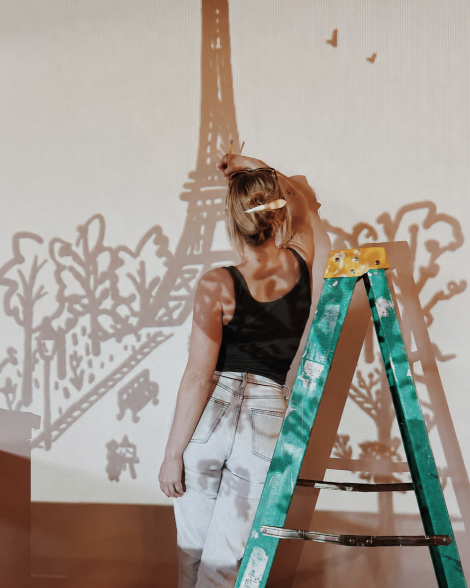 Woman creating a shadow art of the Eiffel Tower and trees on a white wall, using a light source and her hand. She is standing next to a green and yellow step ladder.