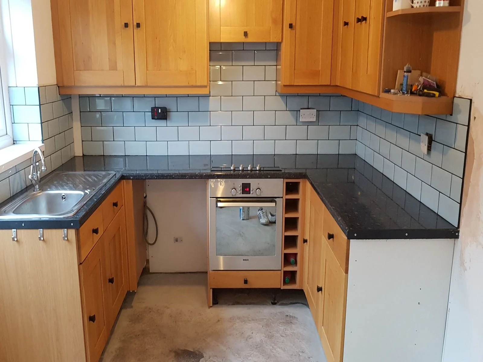 Fitted kitchen installation featuring light oak wooden cabinets, dark granite effect countertops, and light grey metro tiling backsplash.