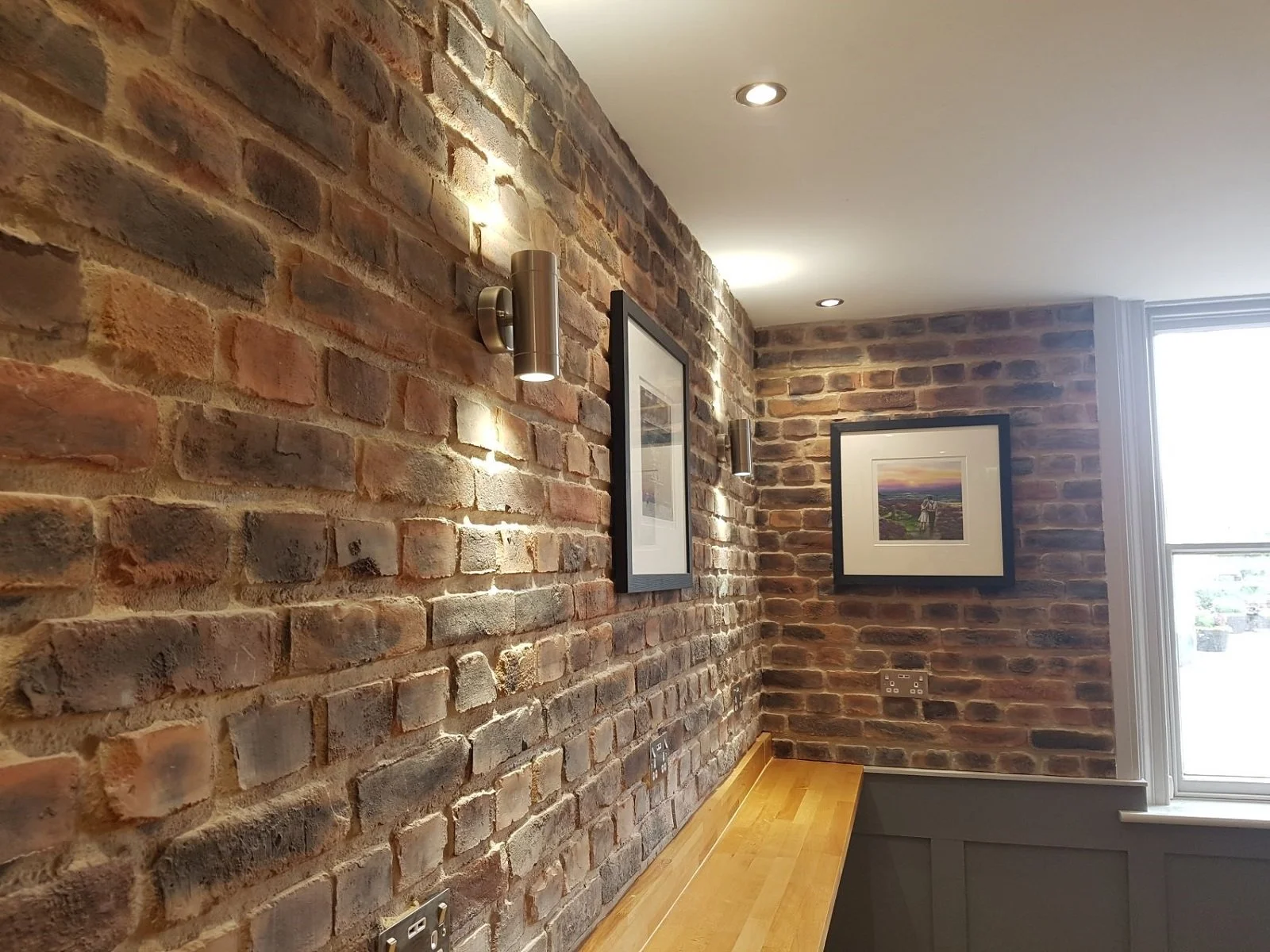 Cozy coffee shop interior refurbishment featuring an exposed rustic brick feature wall accented by modern metallic downlights and framed artwork.
