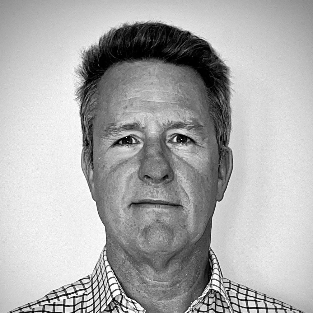 Black and white portrait of a middle-aged man with short, dark hair, wearing a checkered shirt, looking directly at the camera with a neutral expression.