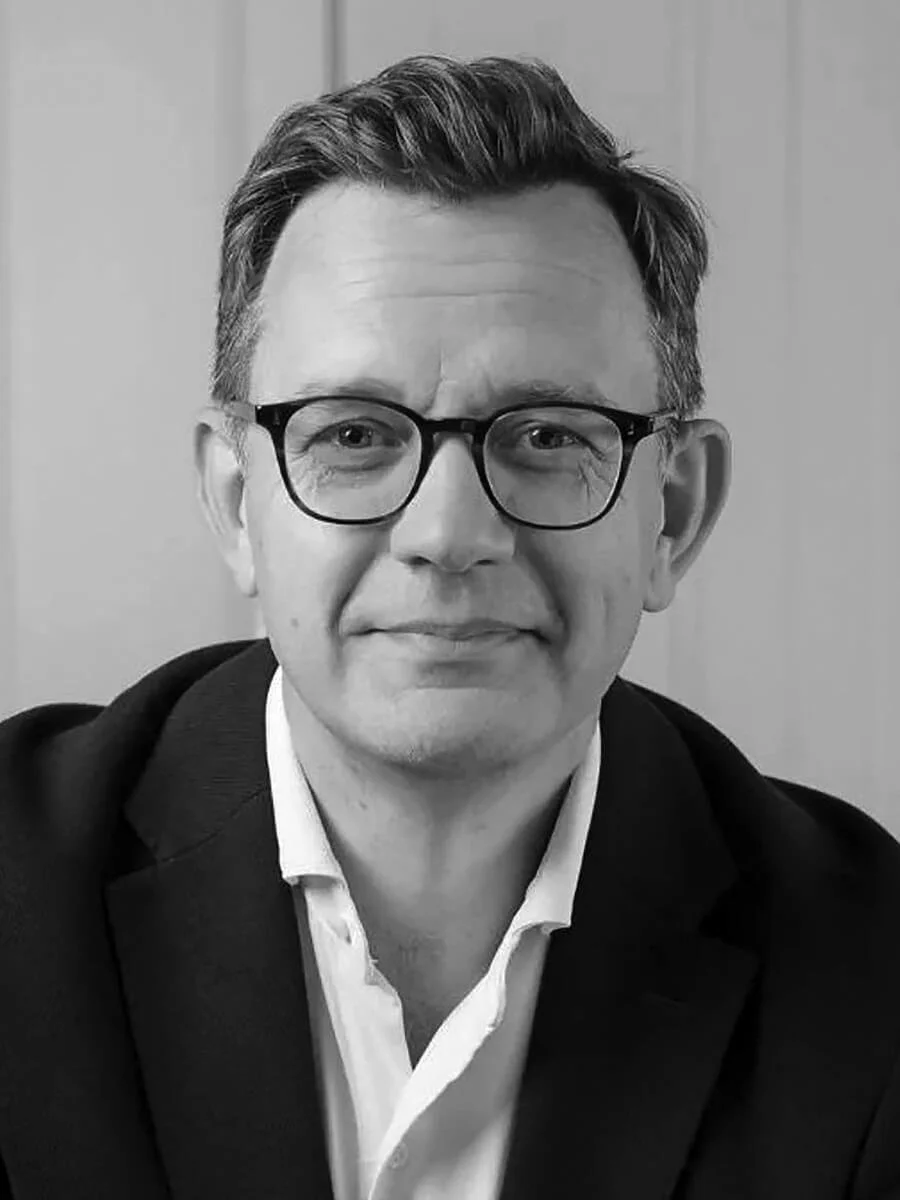 A black-and-white portrait of a man with glasses, styled dark hair, wearing a suit jacket and white dress shirt, smiling slightly against a plain background.