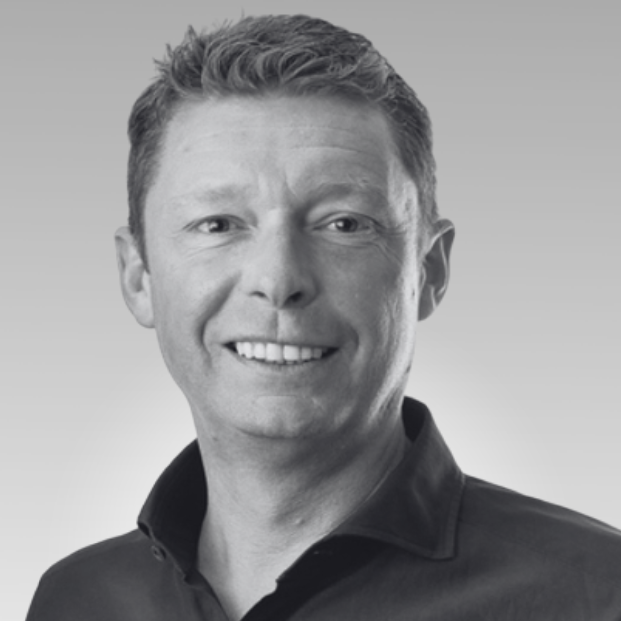 Close-up of a smiling man with styled hair, wearing a dark blazer and shirt against a plain background.