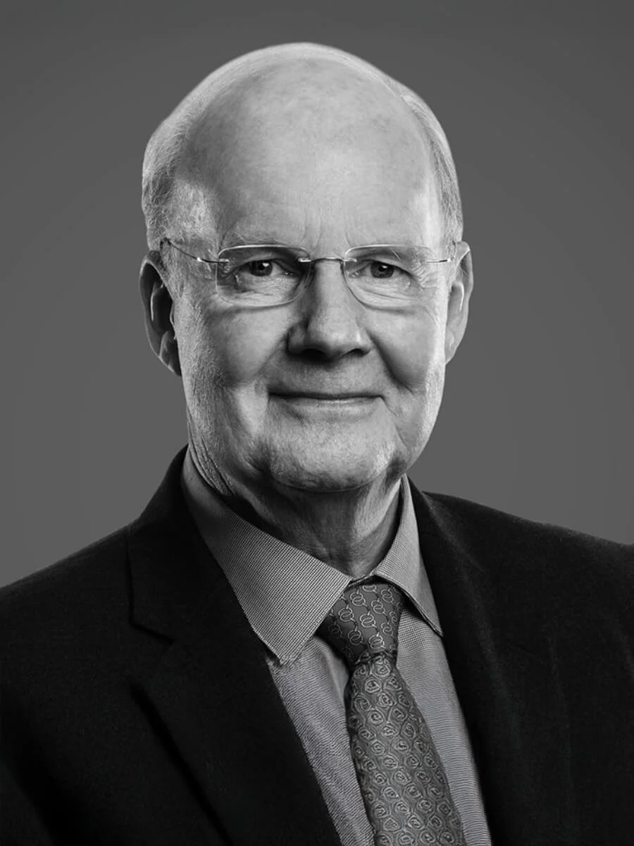 Black and white portrait of an elderly man wearing glasses, a suit, a collared shirt, and a patterned tie, smiling slightly against a plain gray background.