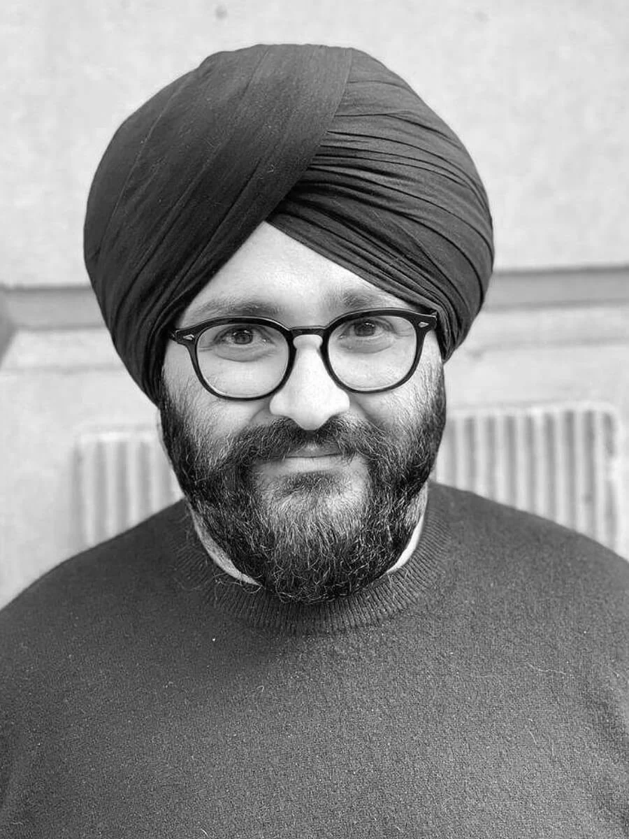 A portrait of a man with a beard and glasses, wearing a turban, smiling at the camera. The photo is in black and white.