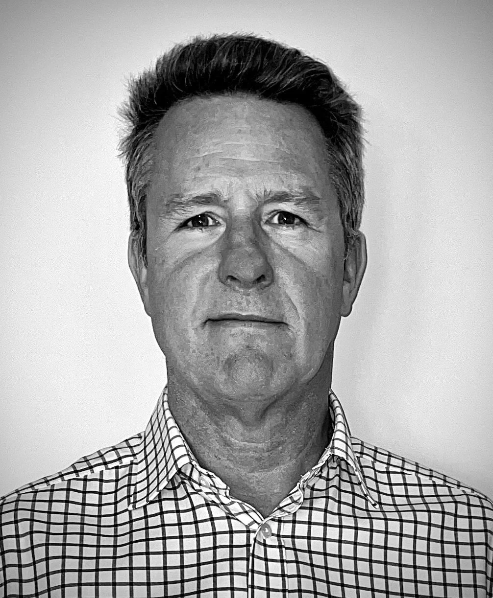 Black and white portrait of a middle-aged man with short, dark and gray hair, wearing a checkered collared shirt, looking directly at the camera with a neutral expression.