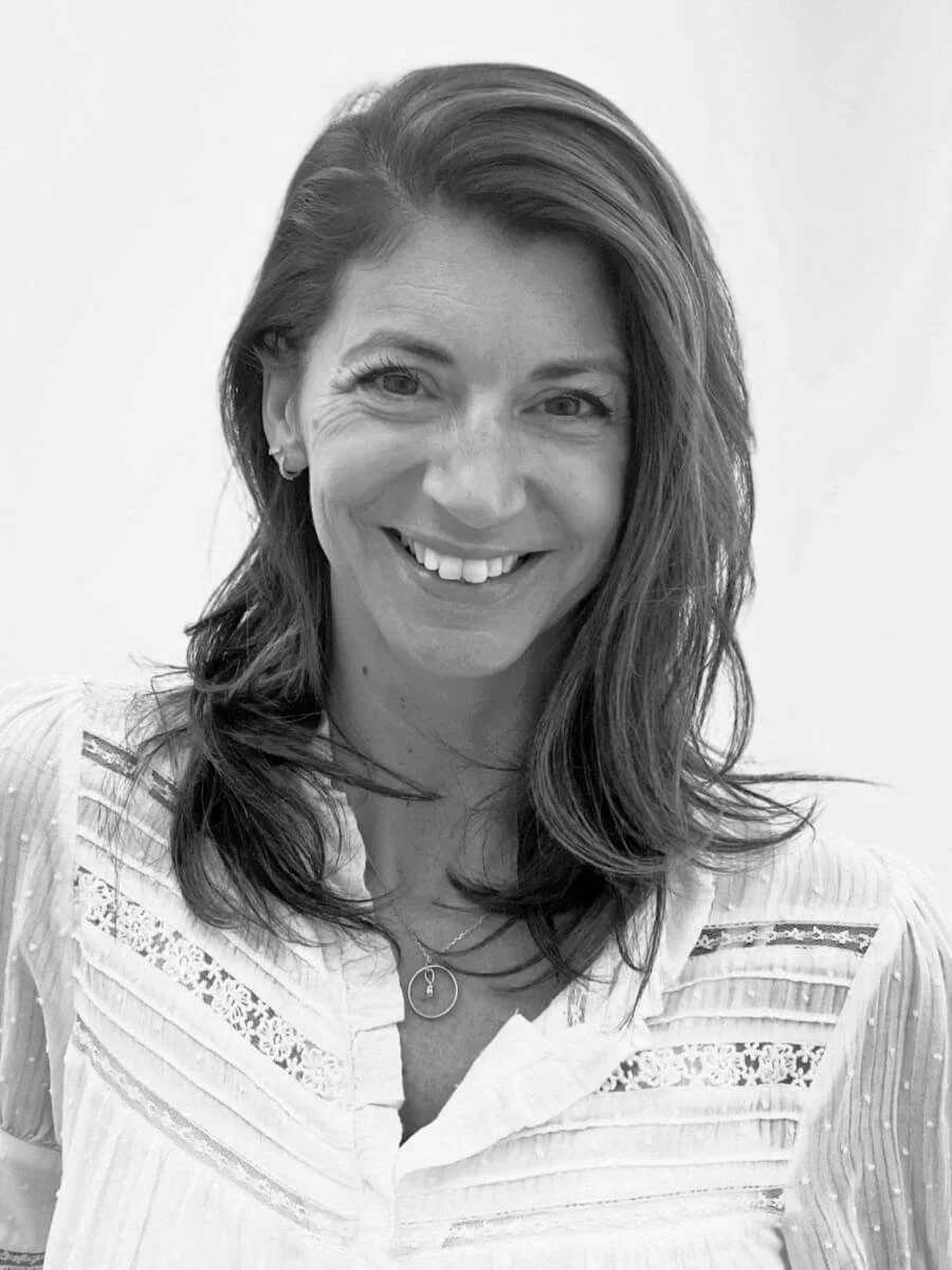 Black and white photo of a smiling woman with shoulder-length hair, wearing a patterned blouse and a necklace, against a plain background.