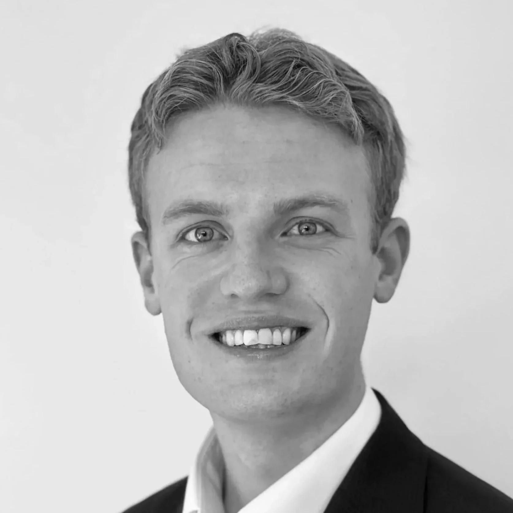 Black and white portrait of a young man with a smile, short wavy hair, dressed in a tuxedo, showing teeth.