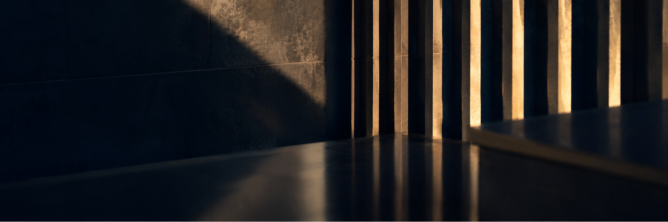 Close-up of a wooden bench and a wall, illuminated by warm sunlight creating shadows.