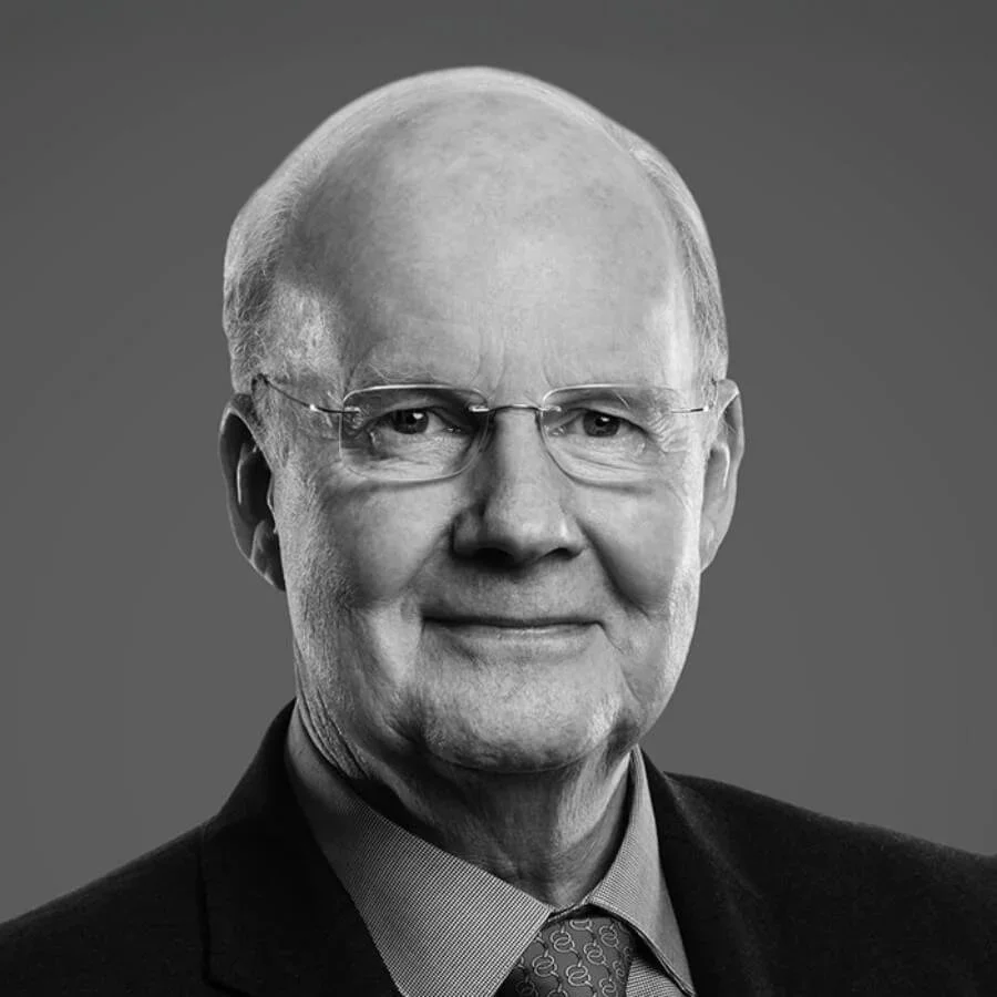 Black and white portrait of an elderly man wearing glasses, a suit, a collared shirt, and a patterned tie, smiling slightly, against a plain background.