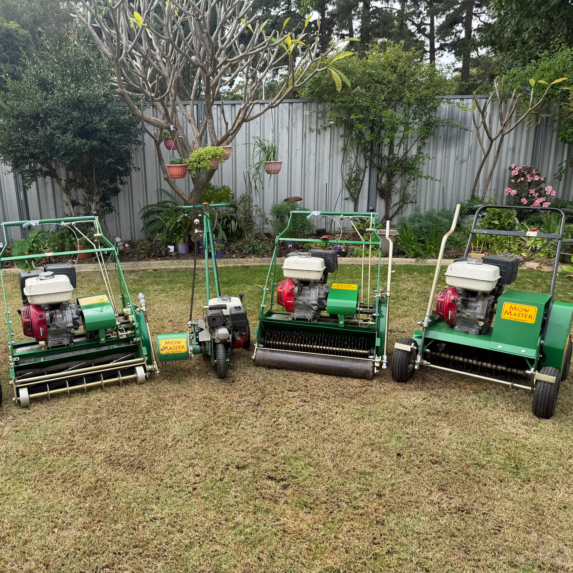 Backyard garden with various trees, plants, and flowers, along with four green and white lawn mowers in the foreground on the grass.
