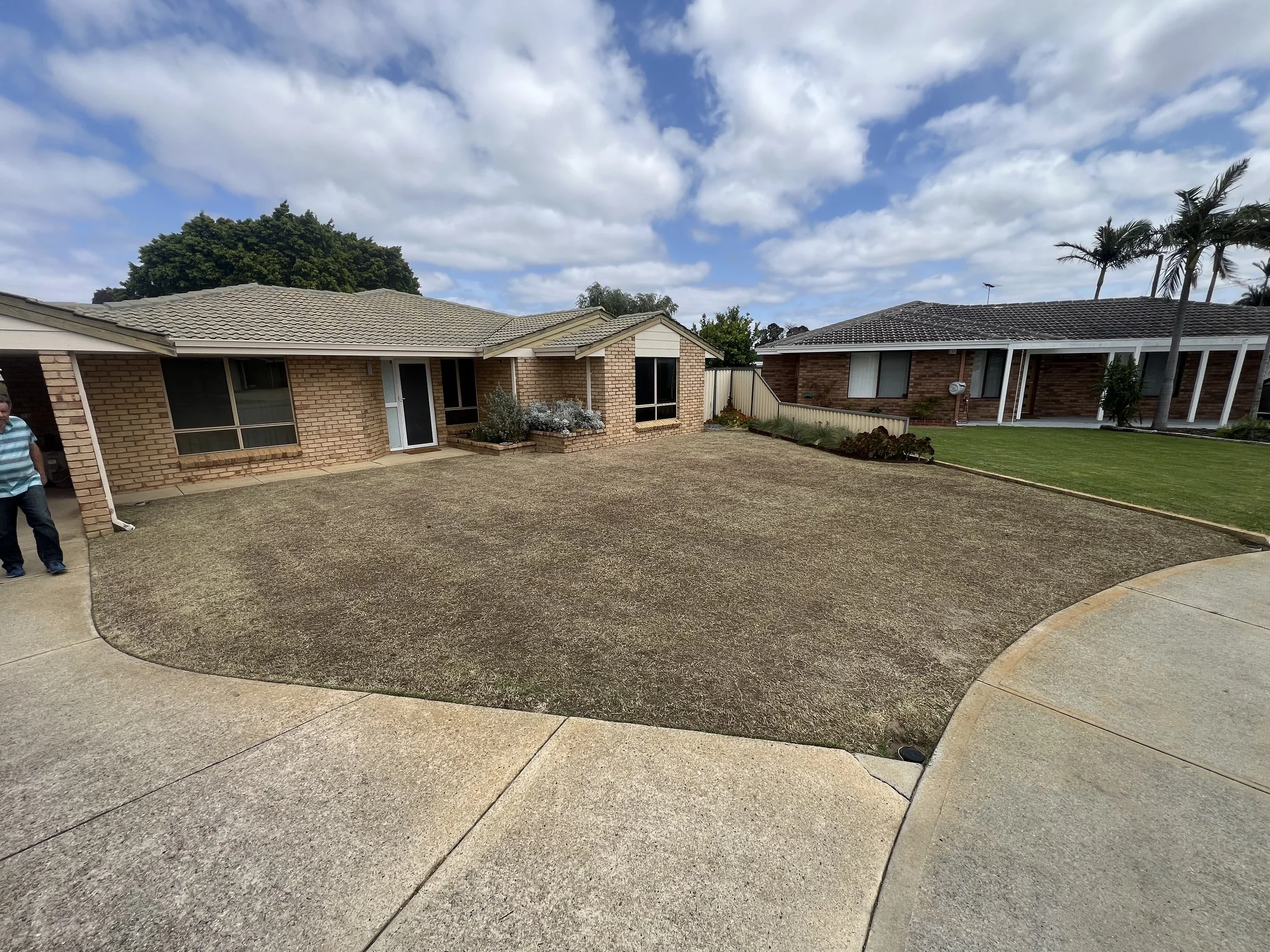 A suburban house with a brick exterior, gray tiled roof, and a small front yard with plants and a walkway, under a partly cloudy sky.