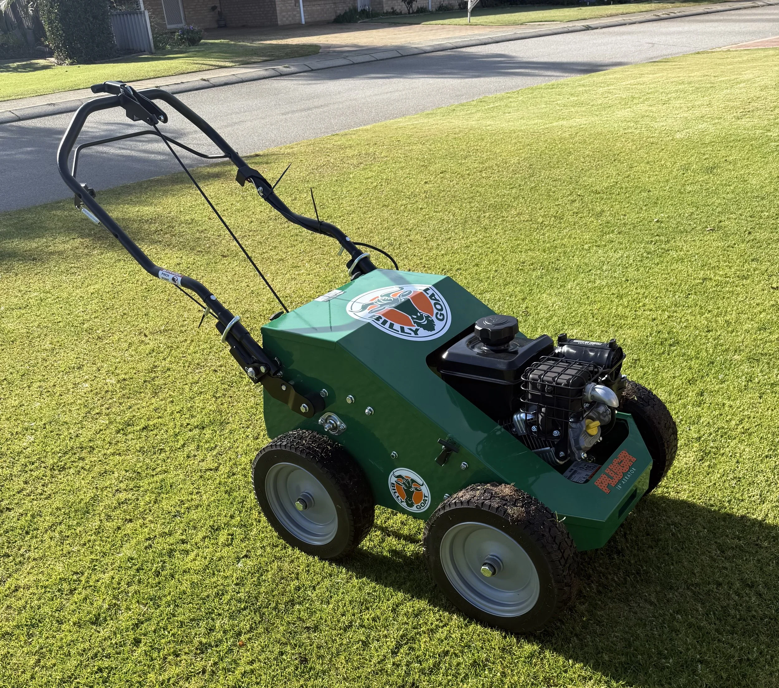 Green lawn mower with a logo on the top, parked on a freshly cut grass yard in a suburban neighborhood, with a sidewalk and a house in the background.