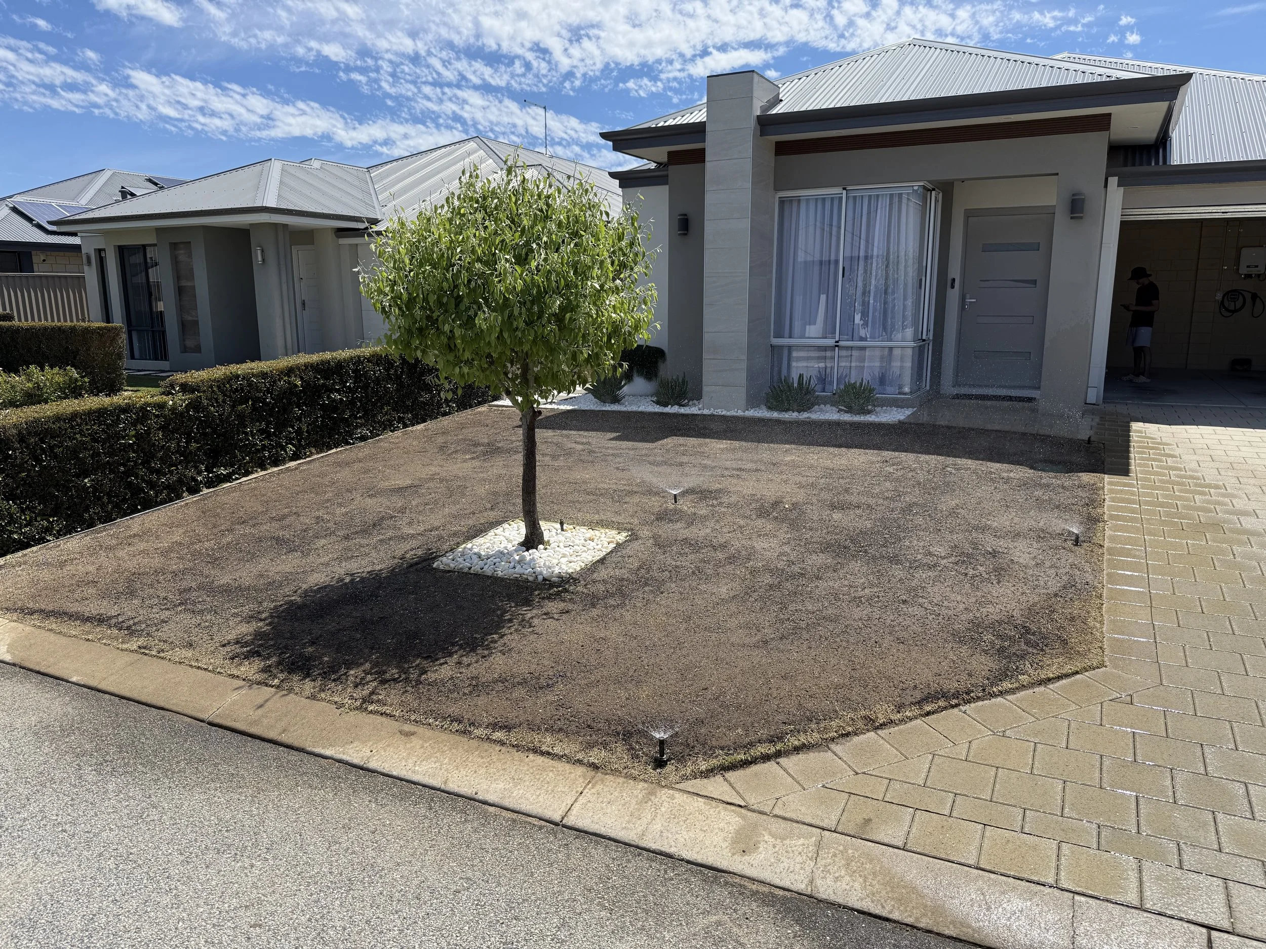 Modern suburban house with grey roof and walls, with a small tree and neatly maintained garden in the front yard, under a partly cloudy blue sky.