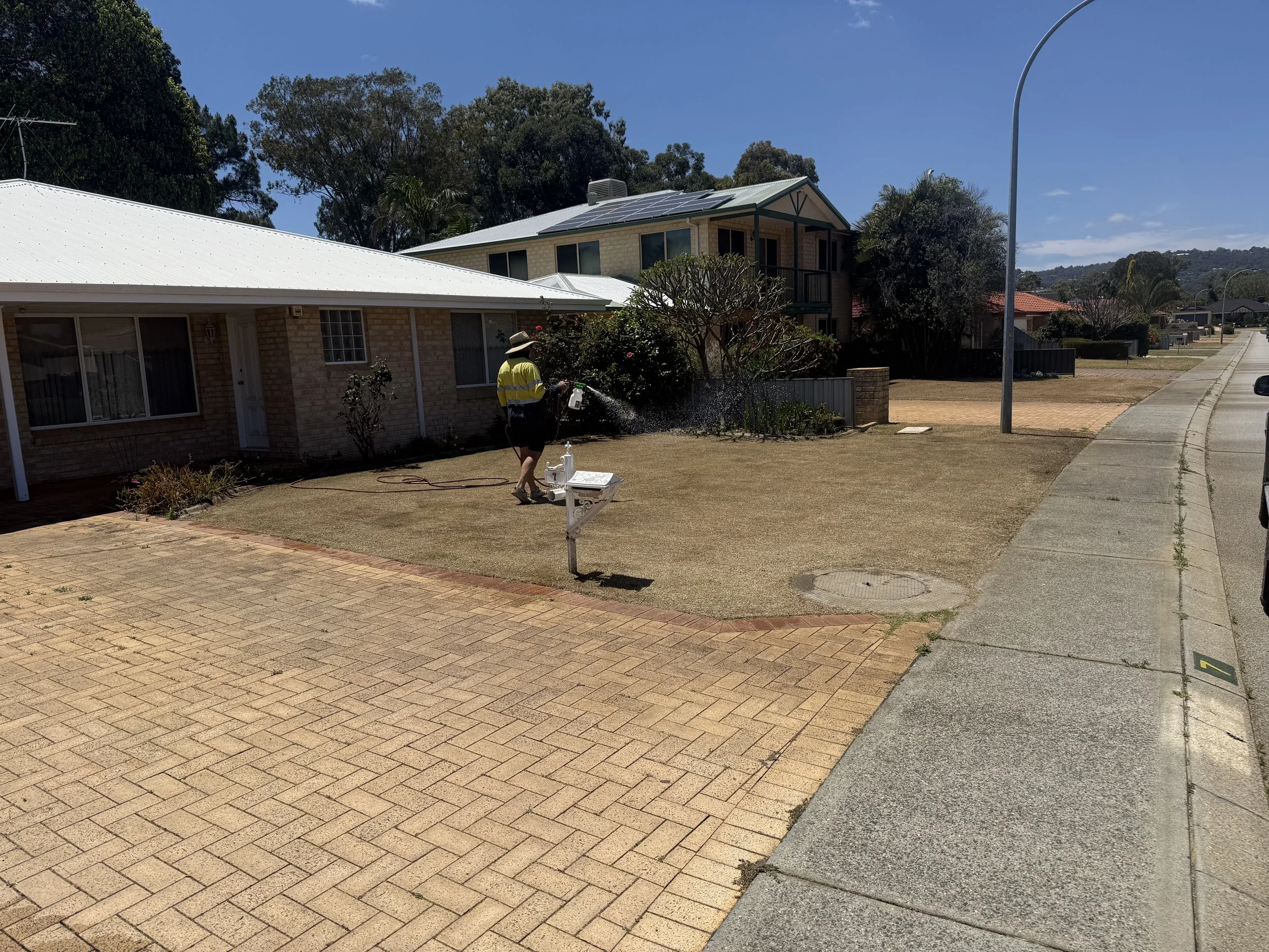 A person wearing a yellow reflective vest and hat is watering the front yard of a house with a garden hose. The house has a brick exterior and a white metal roof. The neighborhood features similar homes with trees and a sidewalk along the street.